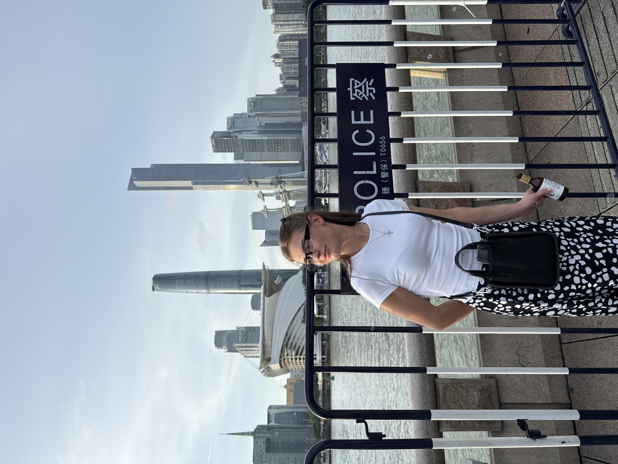 In Haizhu District, China, a young woman with brown hair pulled back and wearing glasses is posing for a photo. She is wearing a white t-shirt and a black and white polka dot skirt. She has a small black purse slung over her shoulder. In her right hand, she is holding a small brown bottle. She is standing behind a black and white metal fence. In the background, there is a body of water and a cityscape with several tall buildings and a stadium. A sign on the fence reads "POLICE" in English and Chinese characters. The sky is blue with a few clouds.