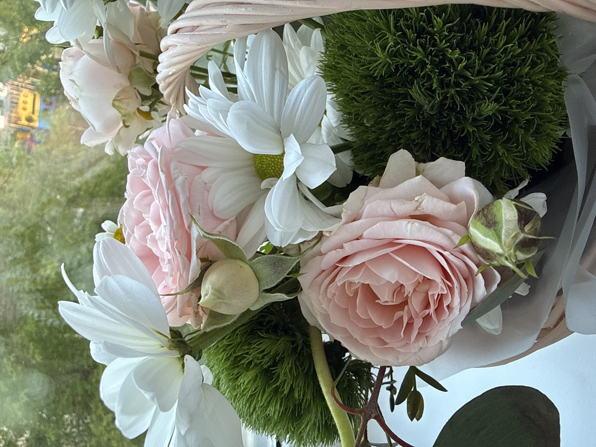 A close-up shot shows a bouquet of flowers in a basket, with light pink roses, white daisies, and green dianthus. A green dianthus is to the right of the bouquet. The arrangement is positioned near a window, with a blurred outdoor background that appears to be in Krasnoyarsk, Russia, including the recognizable bright yellow traffic lights. The flowers are arranged with some white fabric visible around the basket.