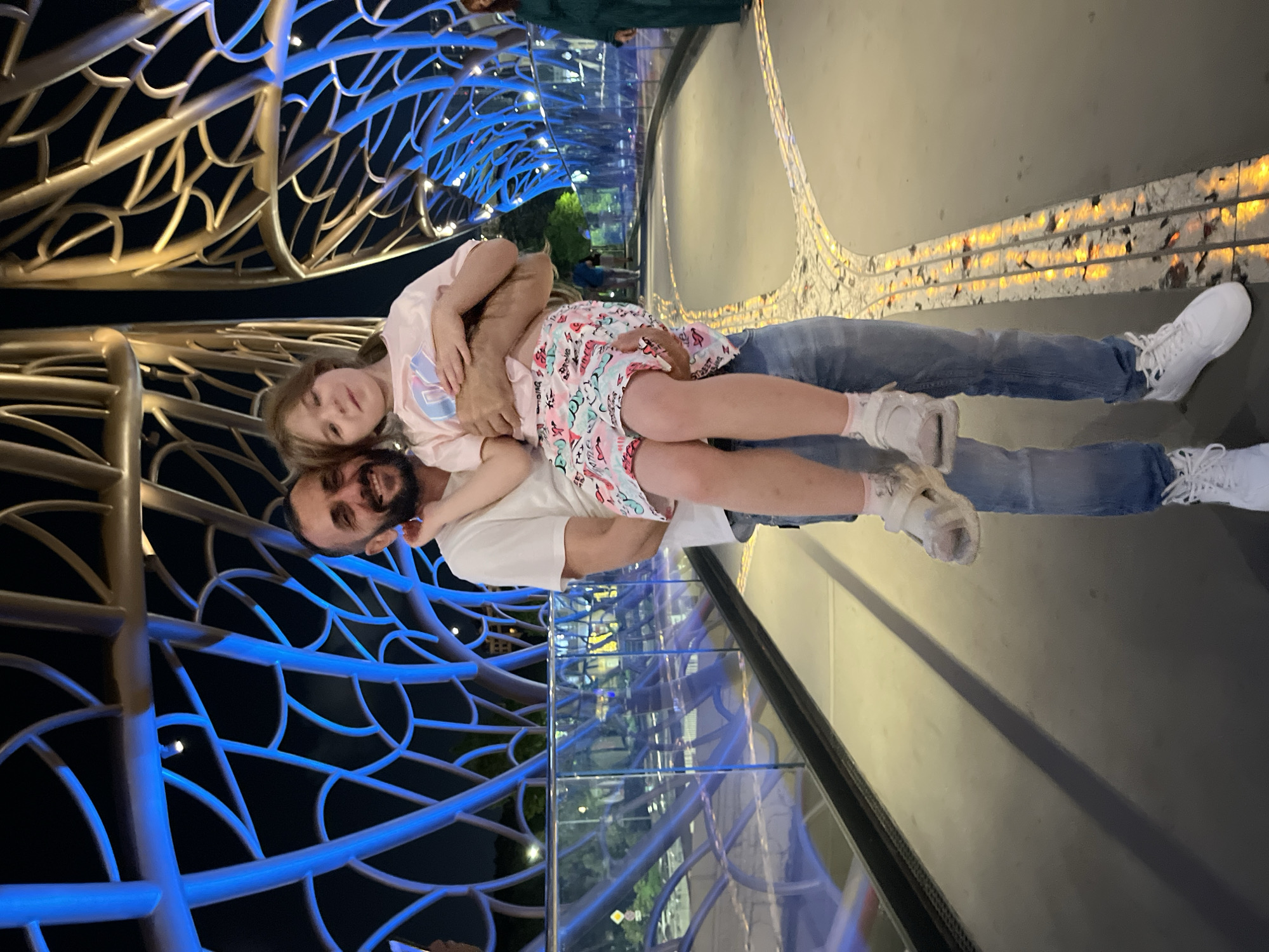 In the city of Sochi, Russia, a man is holding a young girl in his arms on a pedestrian bridge at night. The bridge features a distinctive, sculptural canopy with intertwined metal elements illuminated in blue and gold. The man, with a beard and a white t-shirt, is smiling and looking towards the camera. The girl, wearing a pink t-shirt and a patterned skirt, is also looking at the camera with a slight smile. They are standing on the bridge's walkway, which has a unique mosaic-like pattern embedded with lights. In the background, other people can be seen on the bridge and in the distance, with city lights and trees visible. The overall scene suggests a warm, evening outing.