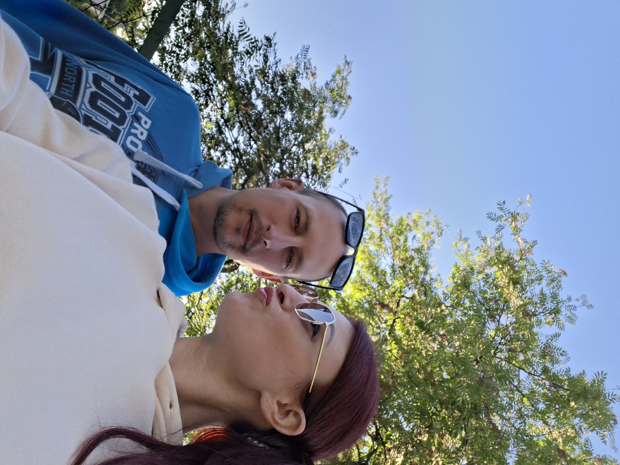 Two people are in the photograph, a man and a woman. The man is wearing a blue hoodie with white writing on the front and has sunglasses perched on his head. The woman is wearing a light-colored sweatshirt and has sunglasses on. The woman has dark red hair. They are standing outside with trees and the sky in the background. The photograph seems to be taken in the city of Забалоцкі сельскі Савет, Belarus.