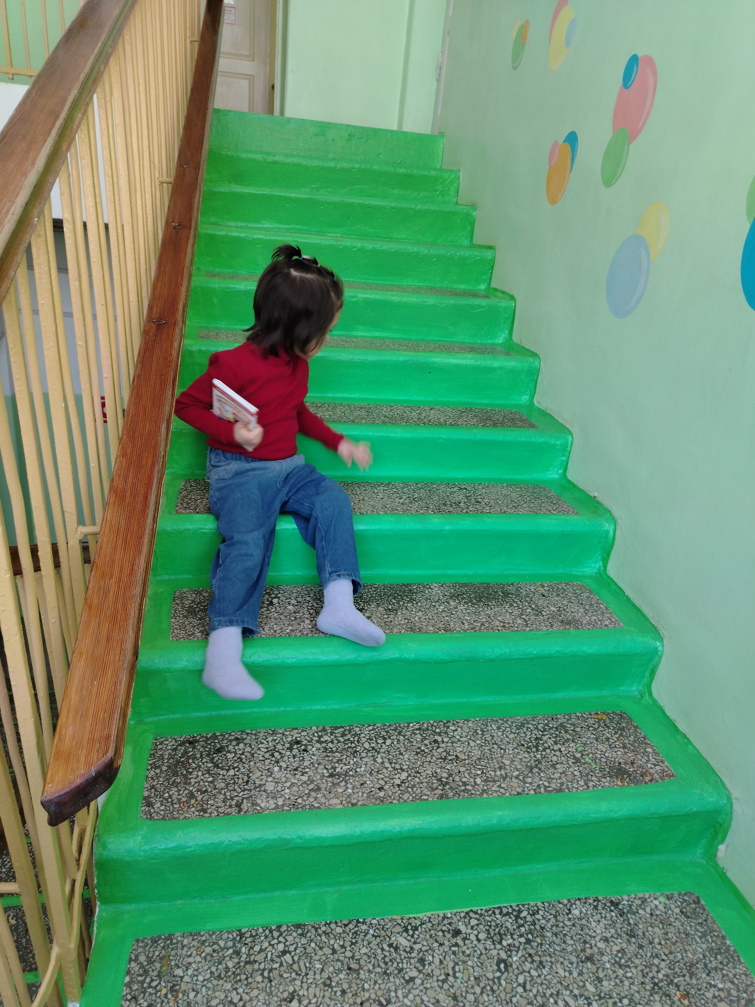 A young child with dark hair sits on a green painted staircase with a pebble aggregate surface. She wears a long-sleeved red shirt, blue jeans, and light blue socks. She holds a book in her left hand and her right hand rests on the stair next to her. The staircase has a wooden banister along the left and a light green wall on the right decorated with colorful oval shapes. The location is in Chaikovsky, Russia.