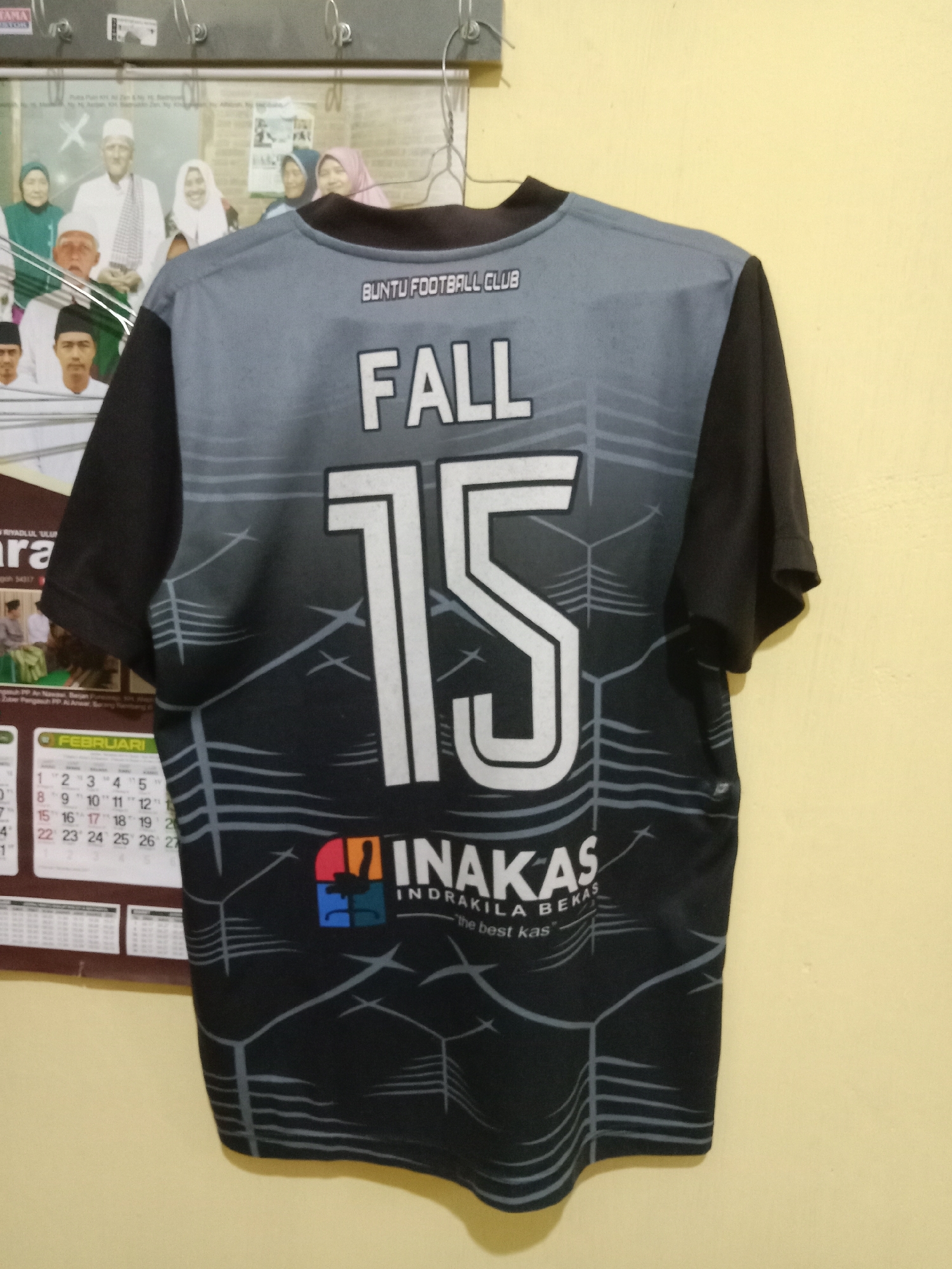 The image shows the back of a black and gray soccer jersey with the number "15" in large white font. Above the number, "FALL" is written in white letters, and above that, "BUNTU FOOTBALL CLUB" is written in smaller white letters. At the bottom of the jersey is a logo with the text "INAKAS INDRAKILA BEKAS" and "the best kas". The jersey is hanging on a wall, and a calendar is visible to the left, showing the month of February. A framed picture or poster with a group of people in traditional attire is also visible behind the jersey. The scene is illuminated by artificial light, casting shadows on the wall. The location is identified as Kutosari, Indonesia.