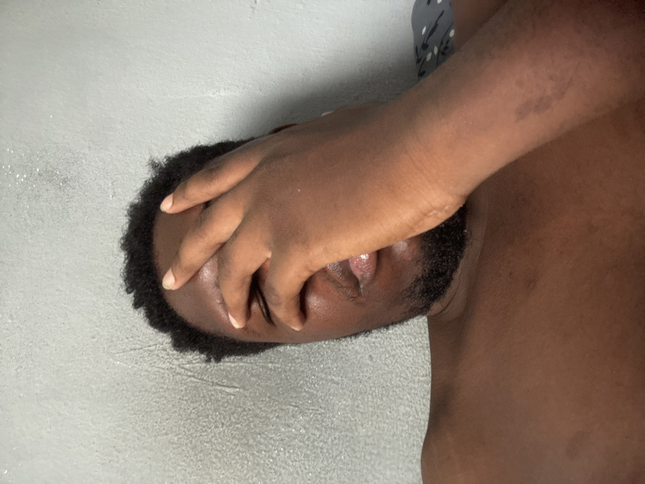 The image displays a male individual with dark skin, short dark hair, and a visible beard, situated indoors in Gbongan, Nigeria. His right hand covers his eyes and forehead. His left shoulder and upper chest are exposed, indicating he is shirtless or partially clothed. The background consists of a light grey textured wall, and a dark fabric with a light patterned design is partially visible behind his right shoulder on the right side of the frame.