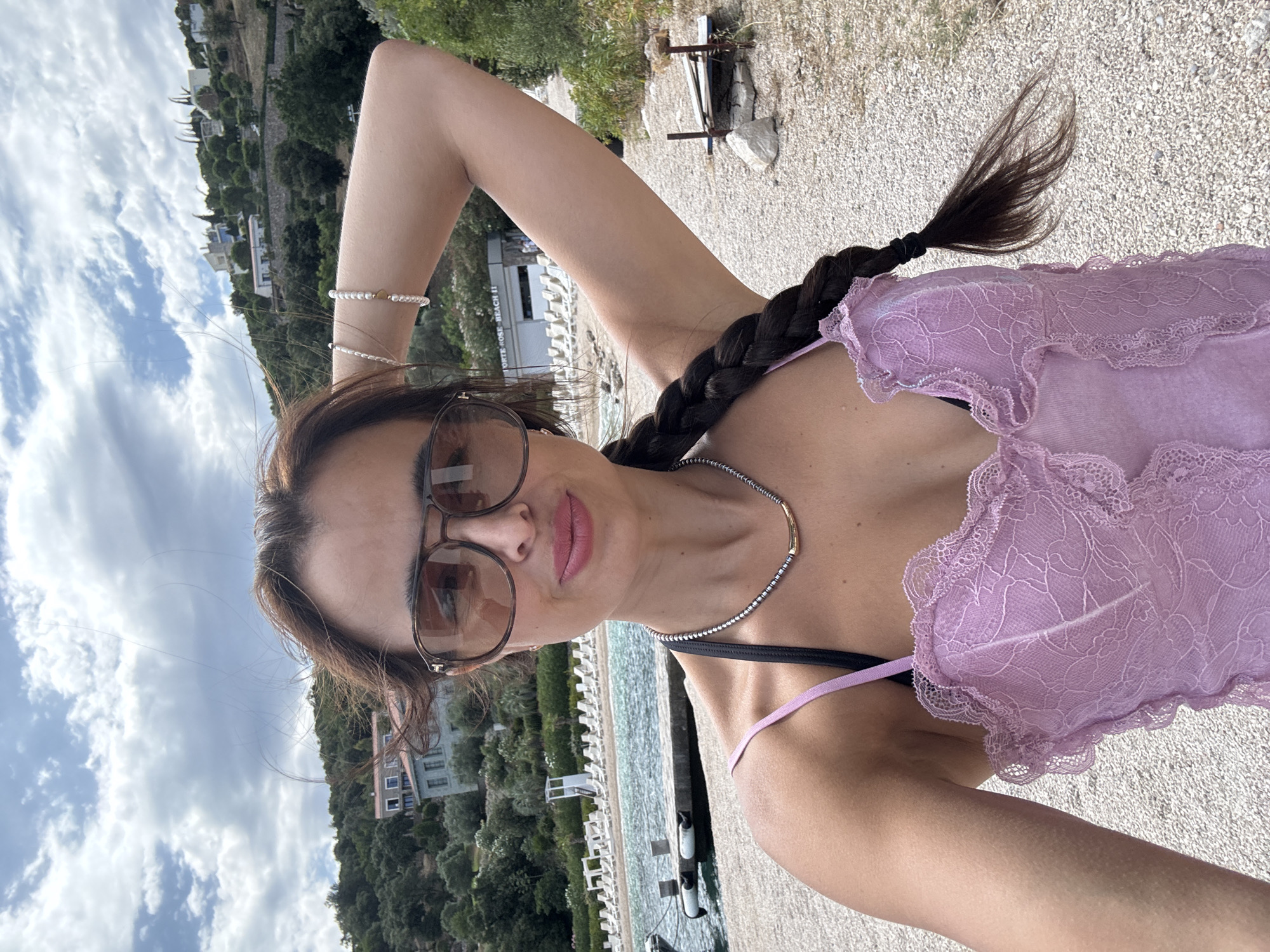 A woman with long, dark hair braided down her back takes a selfie on a pebble beach with a body of water to her left and a hillside dotted with buildings and trees to her right. She is wearing sunglasses, a pearl bracelet, and a light purple lace top with black straps. She has a necklace with a gold pendant. The background shows a beach with rows of lounge chairs, a small dock, and a building with the words "FORTE ROSE BEACH II" visible. The sky is cloudy.