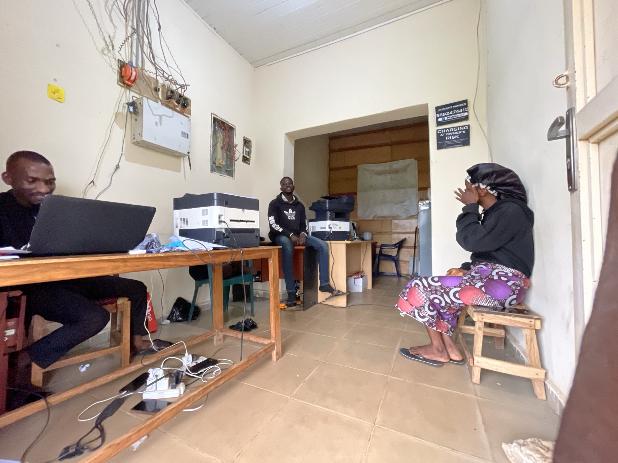The image depicts an indoor scene, likely in Durumi, Nigeria. In the foreground, a man is seated at a wooden desk, using a laptop. The desk is cluttered with electrical cords, a power strip, and several mobile phones charging. To the left of the desk, a printer is visible. In the background, another man is seated on a desk, wearing a black hoodie and jeans. A second printer is positioned behind him. Further to the right, a woman is seated on a small wooden stool, wearing a black headscarf and a colorful patterned skirt. She appears to be looking away from the camera. The room has cream-colored walls with visible electrical wiring and conduits. There are two signs on the wall to the right, one of which reads "ACCOUNT NUMBER 5892474415 Montepoint" and the other "CHARGING AT OWNER'S RISK." The flooring is tiled with a terracotta-like color. The overall environment suggests a functional space, possibly an office or a small business, with a focus on electronic services.