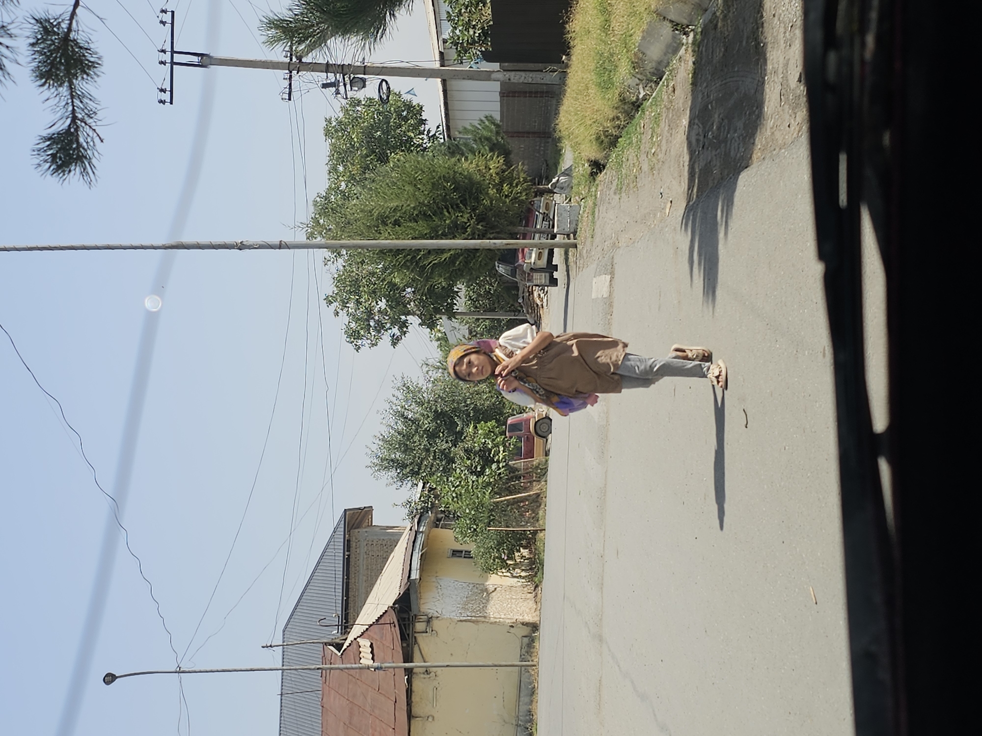 A young girl with a headscarf is walking across a paved road in the city of Özgön, Kyrgyzstan. She is wearing a brown dress over gray pants and light-colored sandals. She is carrying a purple bag on her left shoulder. The girl is the central subject in the image, positioned slightly to the right of the frame. In the background, there is a light yellow building with a brown roof on the left, and a row of trees and a fence on the right. A small red car is parked on the side of the road behind the girl. A street light and utility poles are visible in the background, along with overhead wires. The sky is clear and blue. The image appears to have been taken from a vehicle, as the bottom of the windshield is visible in the foreground.