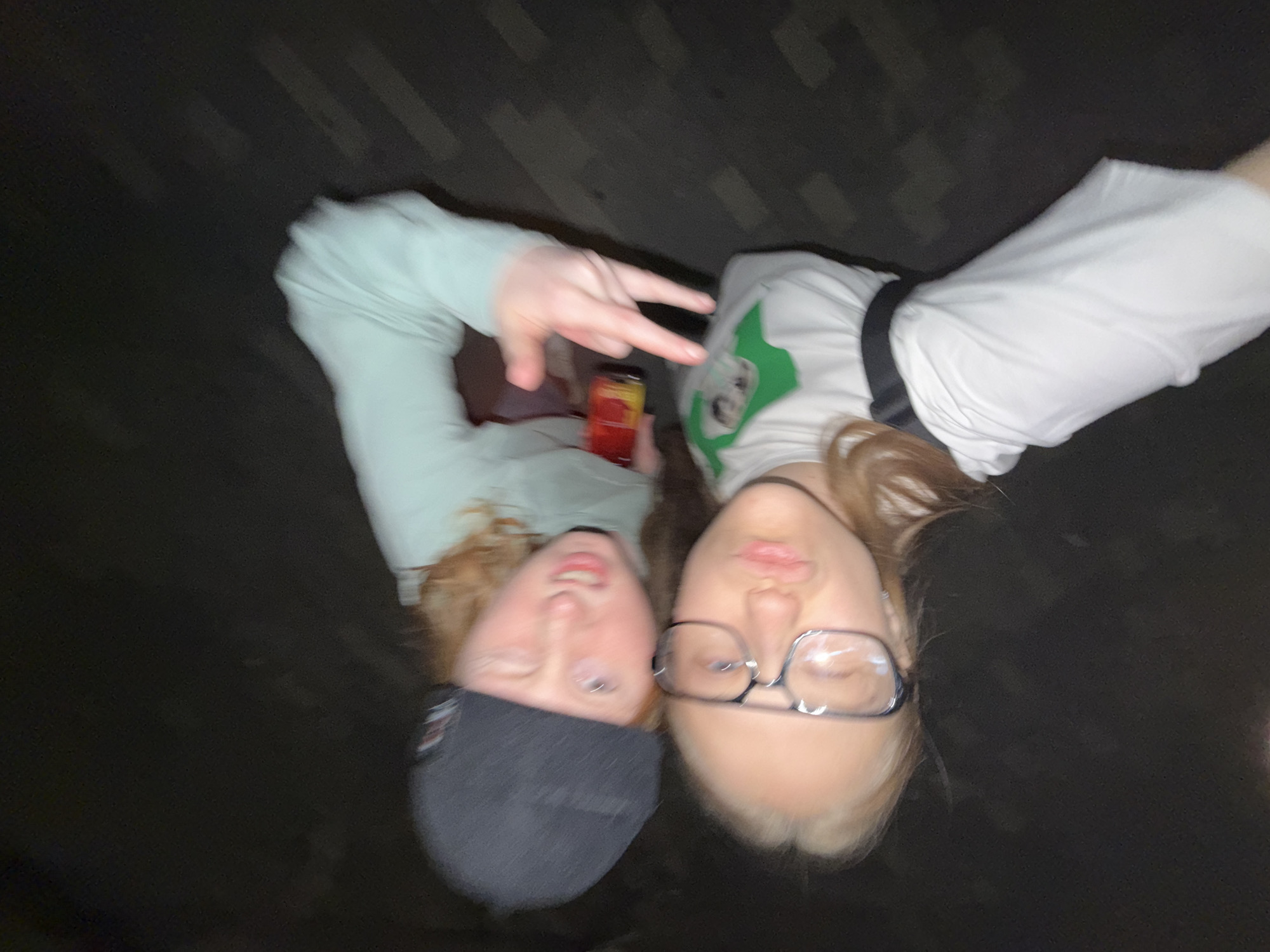Two young people pose for a selfie. The individual on the left wears a grey beanie and a light green long-sleeve shirt, holding a small red object and making a peace sign. The person on the right wears glasses and a white T-shirt with a green graphic. The background suggests an outdoor location at night. There is no information in the picture to suggest that it was taken in Perm, Russia.