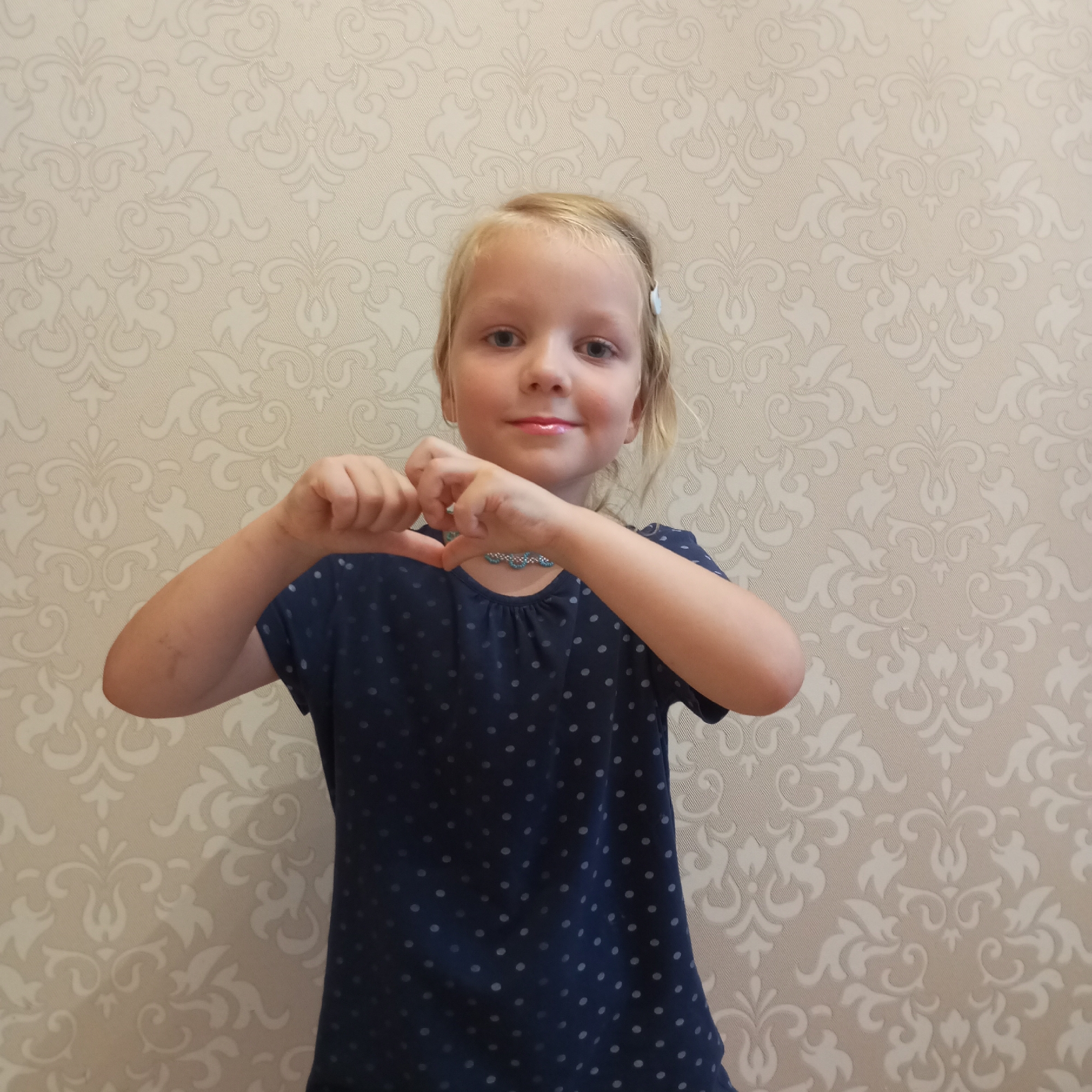 A young Caucasian girl with blonde hair and blue eyes is shown from the chest up, standing in front of a patterned wallpaper. She is wearing a dark blue, polka-dotted shirt and a beaded necklace. Her hands are held together in front of her chest, forming a heart shape. The background is a light-colored wallpaper with a subtle damask pattern. There is no indication of the city of Sochi, Russia, in the image.