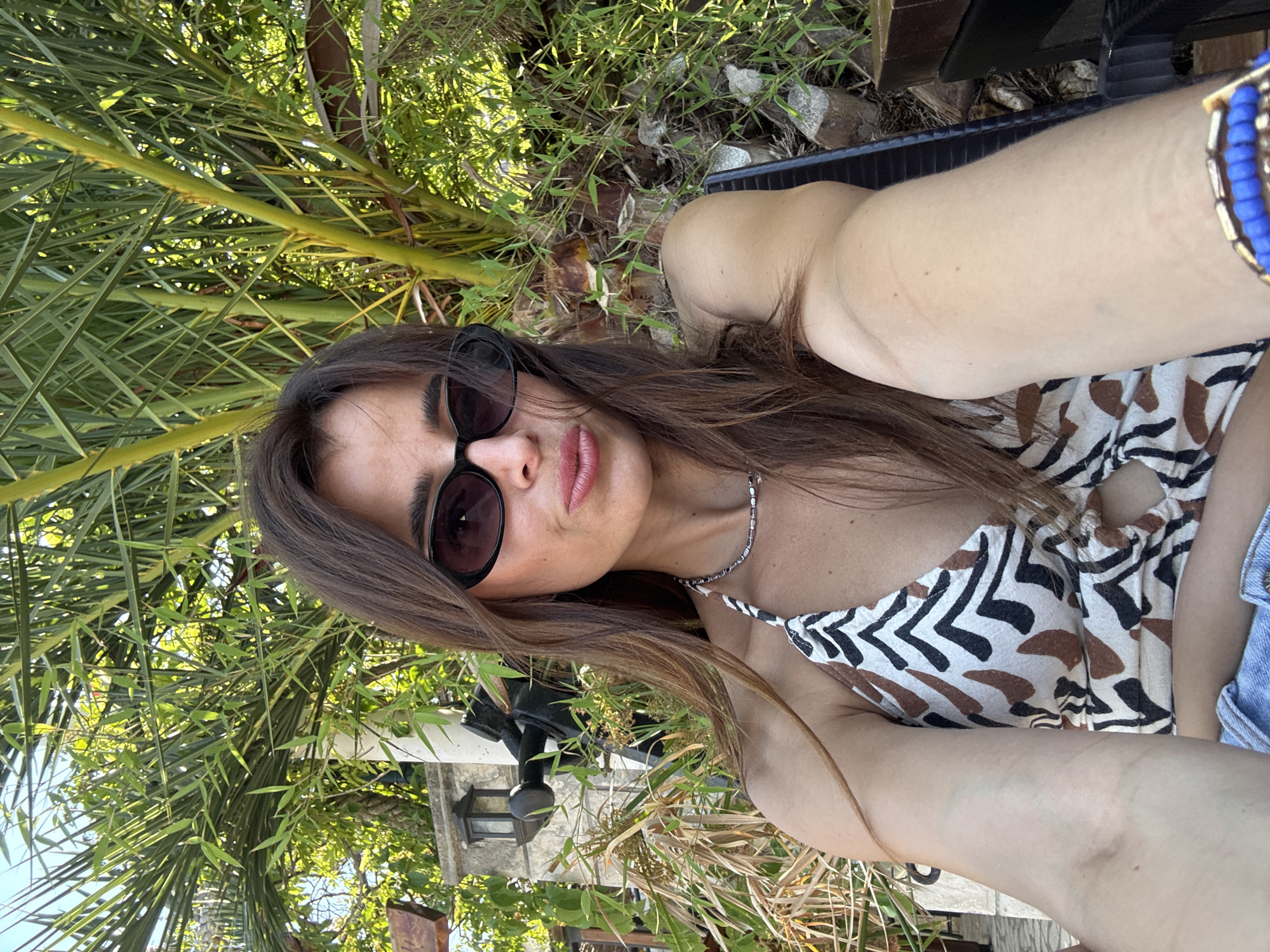 A woman with long brown hair, wearing sunglasses and a patterned top, takes a selfie outdoors. She is smiling with her lips slightly parted and her head tilted. A blue beaded bracelet is visible on her right wrist. Palm fronds and greenery surround her, suggesting a tropical or garden setting. The lighting indicates it is daytime, likely sunny. The background shows some wooden structures and what appears to be outdoor furniture. The image is taken from a slightly elevated angle, looking down at the woman.