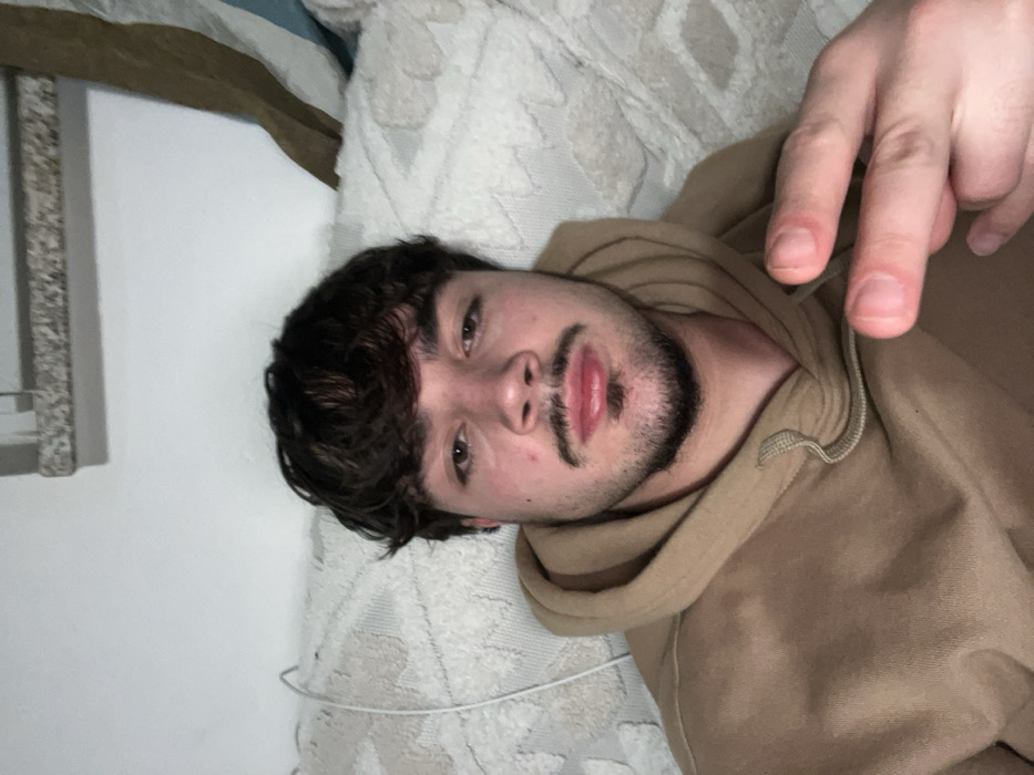 A fair-skinned person with dark curly hair and a trimmed beard lies on a bed. The person is wearing a tan hoodie. One hand is raised, displaying two fingers. The bed has a textured white and beige patterned comforter. A white wall is visible, with a framed object hanging on it. The image appears to be taken at an angle, with the person's head slightly tilted.