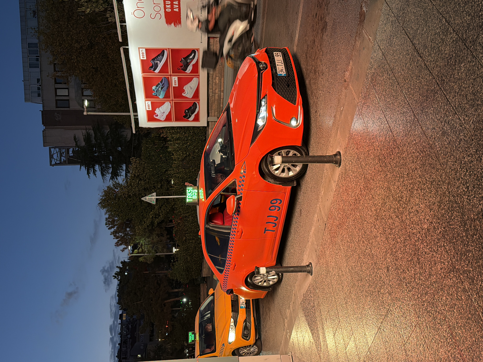 In Kadıköy, Turkey, at dusk, a bright red Toyota Corolla taxi with the license plate "34 TJJ 99" is prominently displayed, parked on a street. The taxi has a distinctive checkered pattern along its side and a green "TAKSİ" light on its roof. Behind it, an orange Fiat Egea taxi, also with a "TAKSİ" light, is partially visible. The street is paved with small stones, and a sidewalk with similar paving is illuminated by warm lighting. In the background, a large billboard advertises shoes with prices in Turkish Lira. A blurred motorcycle with a rider passes by on the right side of the frame. The sky is a deep blue, indicating the transition from day to night, with some clouds present. Trees and buildings are visible in the distance.
