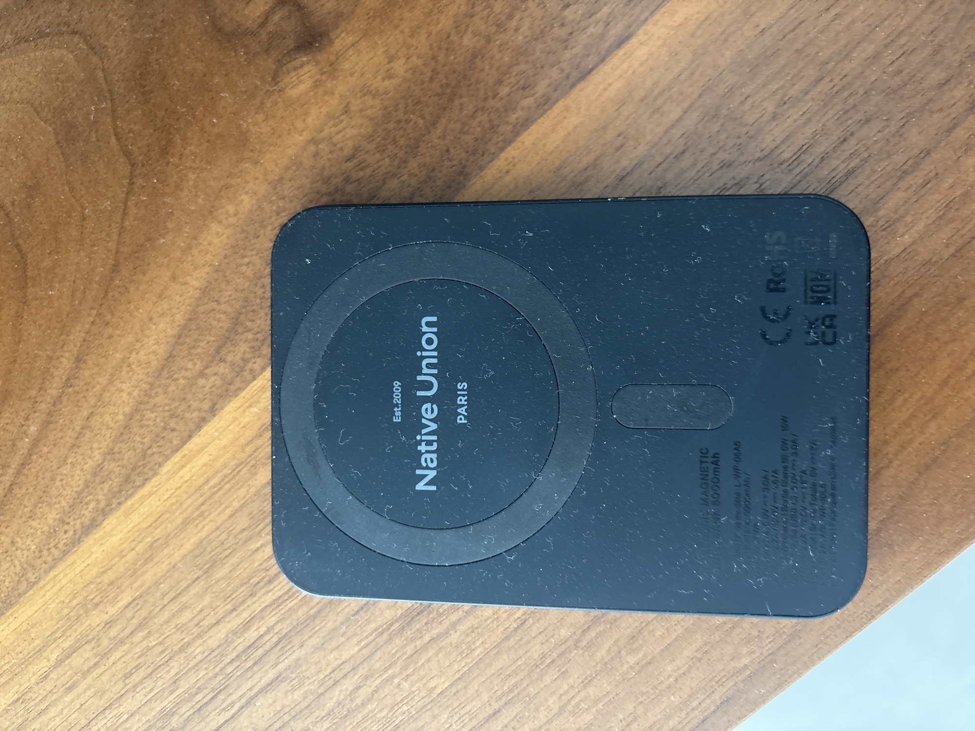 A black, rectangular Native Union magnetic portable charger with a 5000mAh capacity is placed on a wooden table. The charger has brand information and regulatory symbols printed on its surface. The wooden table has a visible wood grain pattern and a warm, brown hue. The lighting suggests an indoor environment. The image is taken from a slightly elevated angle, focusing on the charger and the immediate surface of the table. There is no indication of the city of Dubai, United Arab Emirates in the image.