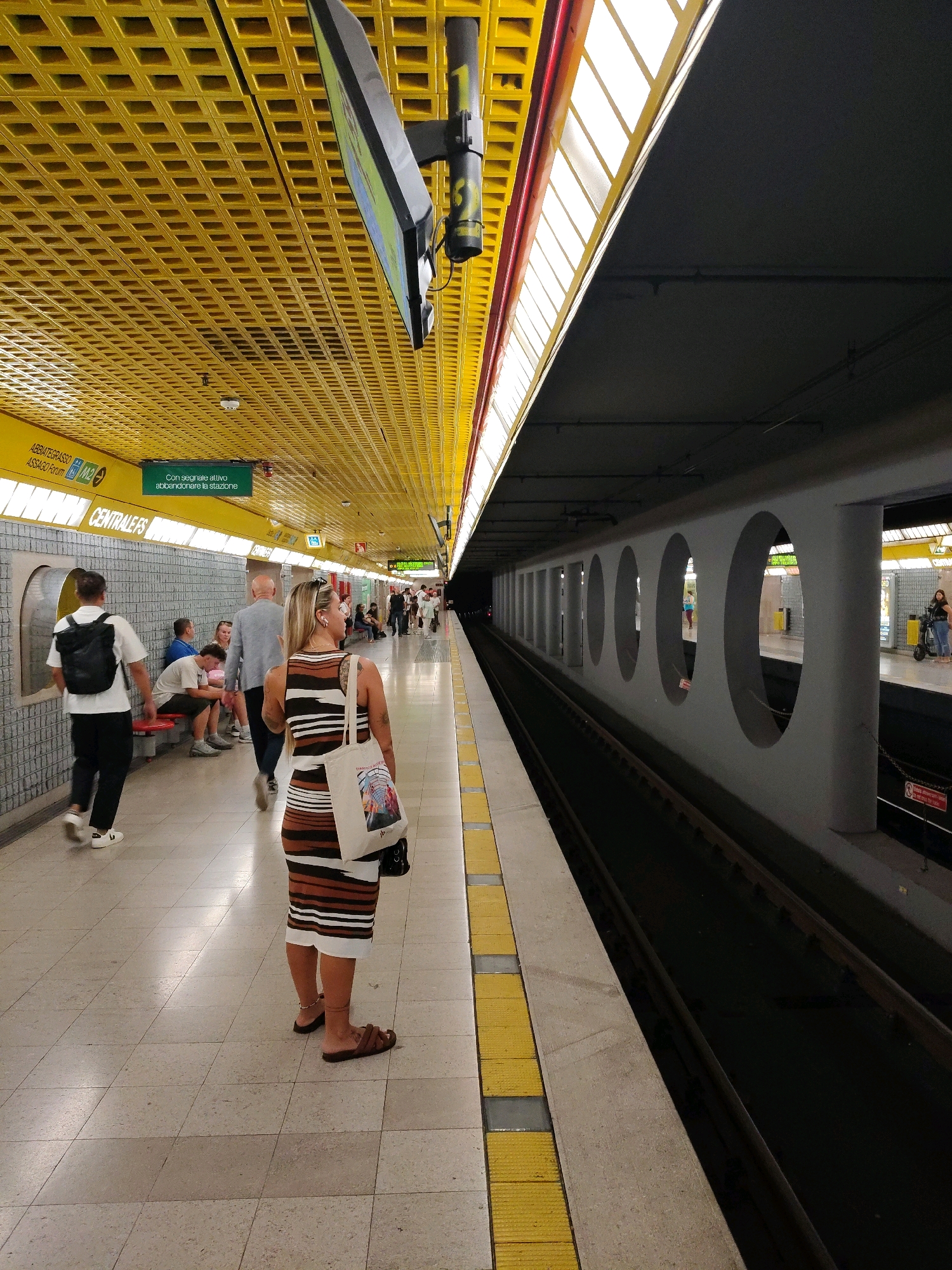 The image depicts a subway station platform in an unspecified urban location. The platform is illuminated by overhead fluorescent lights and features a distinctive yellow, grid-like ceiling. A woman wearing a striped dress and carrying a tote bag stands in the foreground, facing away from the camera. She is wearing brown sandals and has an anklet on her left leg. Other individuals are visible on the platform, some seated on a red bench and others standing. The platform edge is marked with yellow tactile paving. A subway track runs alongside the platform, with a dark, empty tunnel visible in the distance. A gray, circular-patterned wall separates the platform from the track. Signage in green and white is present on the ceiling and walls, indicating station names and directions, though the text is partially obscured or unreadable. The overall scene conveys a sense of urban transit and everyday activity.