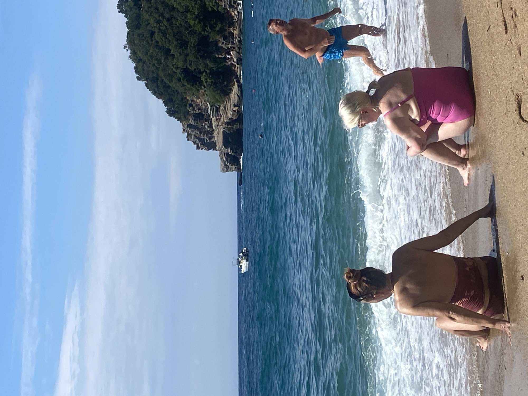 This image depicts a vibrant coastal scene at Sveti Stefan, Montenegro, under bright daylight. The setting features a sandy beach leading into the clear blue sea, with a dramatic, tree-covered rocky cliff rising from the water on the left side of the frame. The sky is a clear, light blue with minimal, wispy clouds.
Several individuals are enjoying the beach and water. In the mid-ground, a man with light hair, wearing blue patterned swim shorts, stands partially submerged in the shallow, wavy water, facing towards the shore. Closer to the foreground on the sandy beach, two women are crouching at the water's edge. One woman, with blonde hair, wears a pink one-piece swimsuit, while further down the beach, another woman with dark hair styled in a bun wears a dark red two-piece swimsuit. Both appear to be relaxing by the gentle waves lapping the shore.
In the distance, a small white boat with people onboard is visible on the calm, open water. The overall impression is one of a serene and leisurely day spent at a picturesque coastal location. There is no visible text in the image.