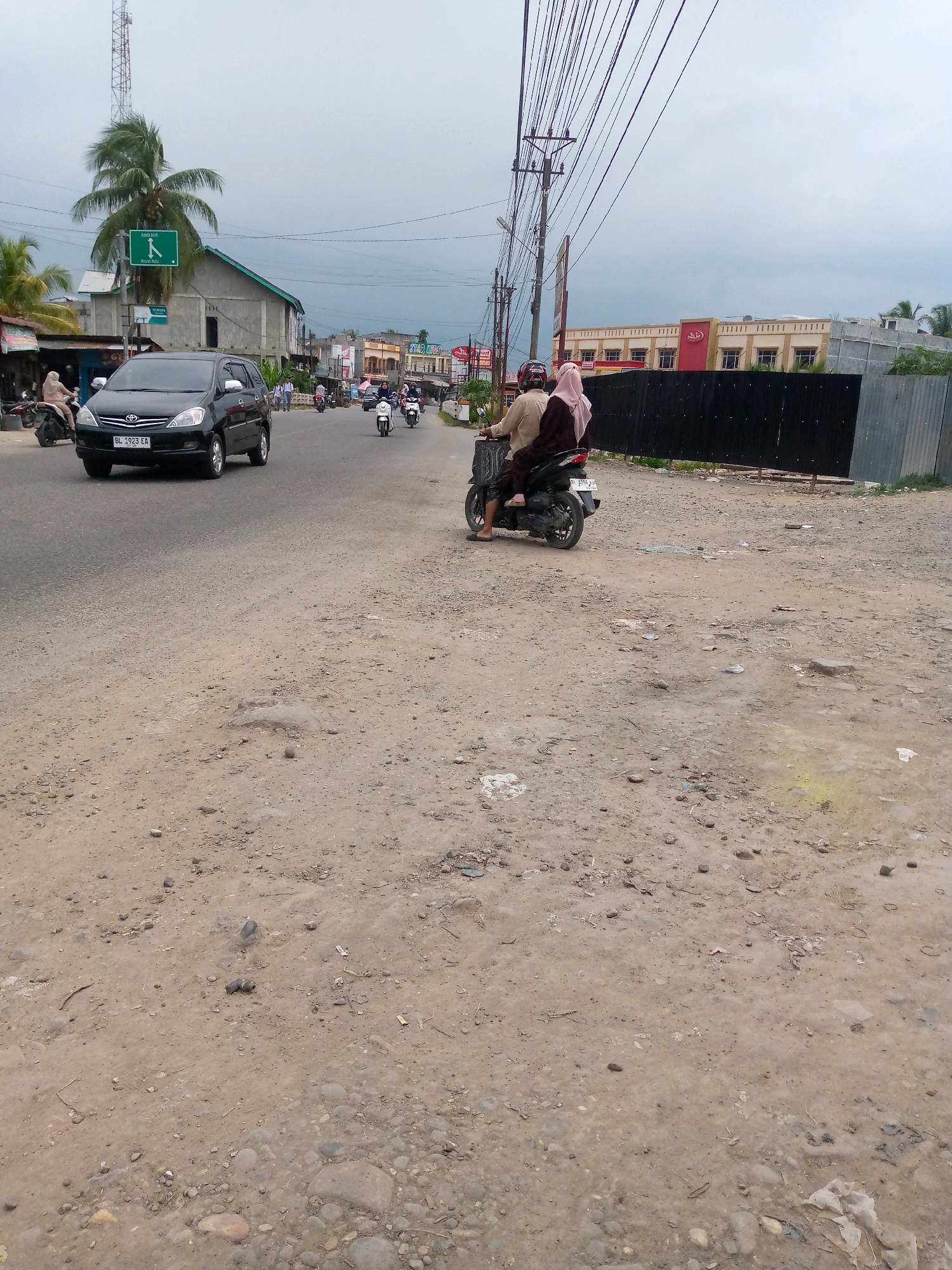 The image shows a street scene in Keudegeudong, Indonesia. A black Toyota Innova with license plate BL 1923 EA is traveling on the asphalt road. To the right of the car, a couple rides a motorcycle, the driver wearing a helmet and the passenger a headscarf. Further down the road, several other motorcycles are visible. Buildings line the street, including a structure with a sign that says "STAR HOTEL." The ground beside the road is unpaved and appears to be dirt with scattered gravel and some puddles of water. Overhead, power lines crisscross the sky, which is overcast. A tall communication tower with a palm tree beside it is visible in the background.