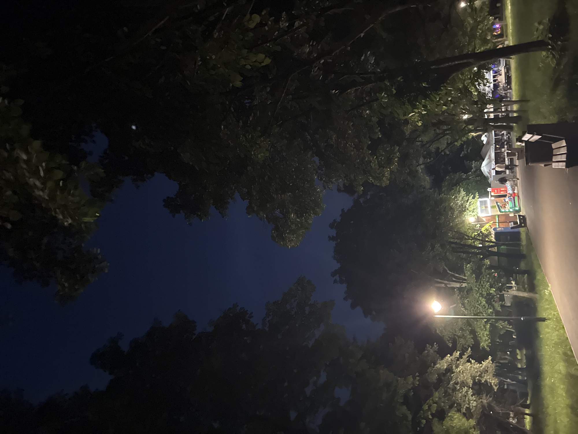 The media file presents an outdoor scene at night, likely in a park or public area. The image is dominated by a dark blue sky, framed by the silhouettes of dense tree canopies on the left, top, and bottom. A bright street lamp with a warm glow stands prominently on the right side of the frame, illuminating its immediate surroundings.
Along the right edge of the image, a paved path or road runs parallel to a grassy area. In the background, beyond the trees and path, several temporary white tents or gazebos are visible, suggesting an evening market, fair, or gathering. Dimly lit figures of people can be seen congregated around these structures, indicating social activity. Further back, objects resembling park benches are present. Near the bottom right, there appears to be a smaller, colorful stand, possibly for food or merchandise, adding to the festive atmosphere.
The time of day is clearly nighttime, evidenced by the dark sky and reliance on artificial light sources. The overall impression is that of a calm evening event in an urban park. No discernible text is visible in the image. Given the context, this scene depicts an evening in Burgas, Bulgaria.