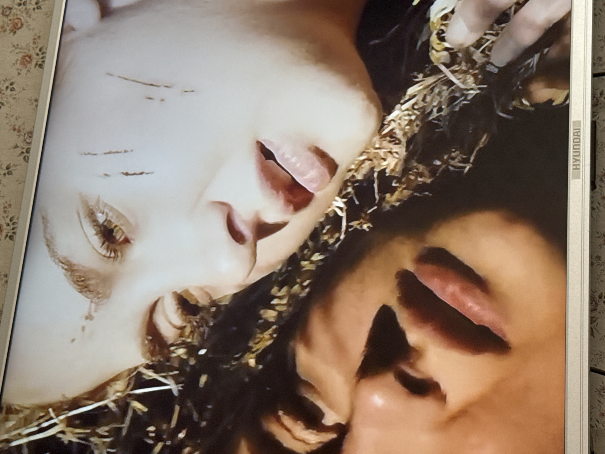 The image shows a close-up of two faces lying next to each other in what appears to be hay or straw. One face has what appear to be scratches on the forehead and a tear streak on the cheek. The Hyundai logo is visible on the side of what seems to be a screen or monitor displaying the image. The image does not provide enough context to determine a specific location in Mytishchi, Russia.