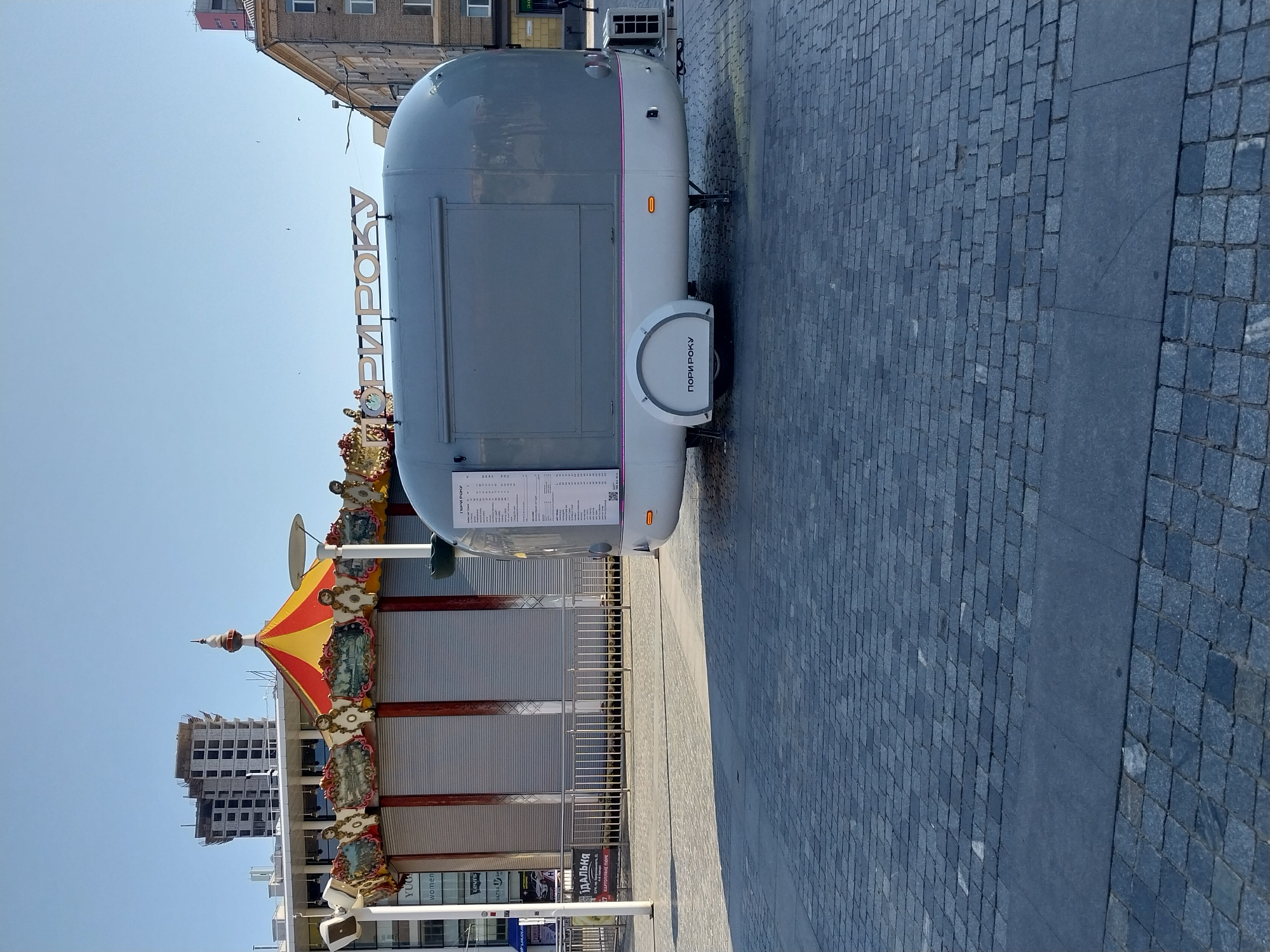 The image depicts an outdoor scene in the city of Dnipro, Ukraine, likely on a sunny day given the clear blue sky and the sharp shadows cast on the cobblestone pavement. In the foreground, a grey, teardrop-shaped food trailer is parked, with a menu displayed on its side. The trailer has "ПОРИ РОКУ" (Porі Roku - Seasons) written on it in both Ukrainian and Russian. Behind the trailer, a carousel with a red and yellow striped canopy and ornate decorations stands closed, with its metal shutters down.
Several buildings are visible in the background. To the left, a modern, multi-story building under construction, with exposed concrete and window frames, dominates the skyline. Further left, the facade of a commercial building is partially visible, with signs for "YUG," "women," and "Levis." To the right of the carousel, a light-colored building with a pink section at the top is visible, and to its right, a brick building with multiple windows. The ground is paved with dark grey cobblestones, arranged in a pattern. A metal railing separates the food trailer and carousel area from the pavement. No people are visible in the image.