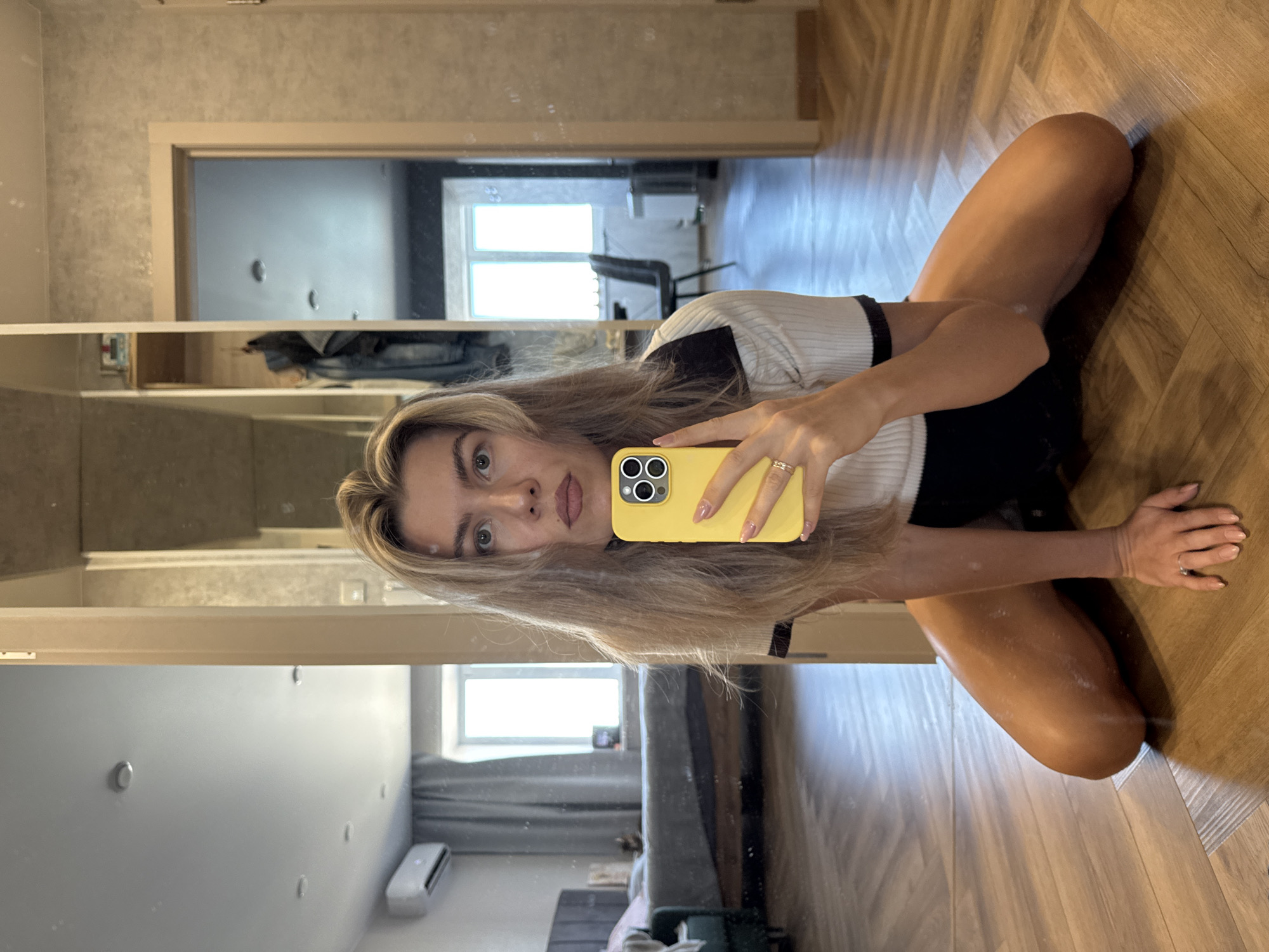 A person is taking a selfie in front of a mirror. The reflection shows a bedroom with a bed, air conditioning unit, and a window. The location is unknown.