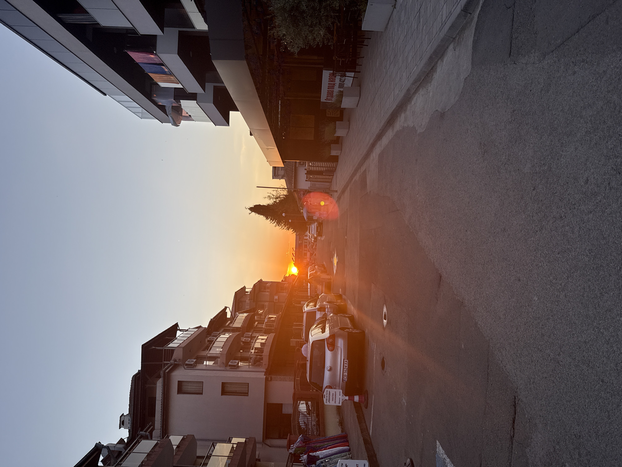 This is a street-level photograph taken in the city of Lozenets, Bulgaria, during sunset. The sun is low on the horizon, casting a warm orange glow across the scene and creating lens flare on the left side of the image. Two multi-story buildings flank a paved street. The building on the left appears to be residential, with balconies and air conditioning units visible. The building on the right has a modern, dark facade and an outdoor seating area with tables and chairs. Several cars are parked along the street. Signs are visible on some of the parked cars and on the building on the right. The sky is clear with a gradient of blue and orange. The pavement is textured, and there is a curb separating the street from a sidewalk made of interlocking bricks.