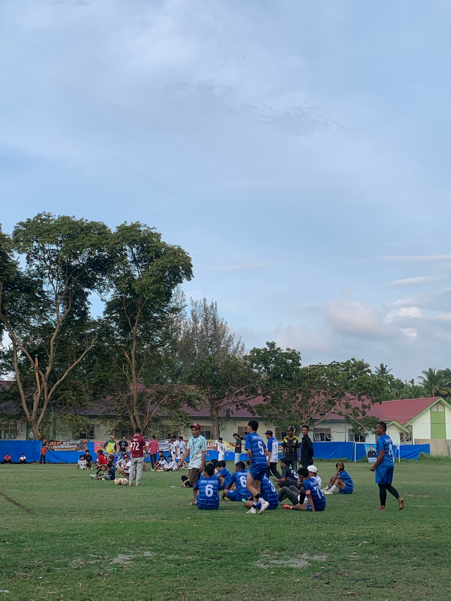The image depicts an outdoor scene on a grassy field, likely a sports ground, in Keudelapang, Indonesia. A group of young men, dressed in soccer uniforms, are scattered across the field, some sitting and others standing. Several individuals are wearing blue jerseys with numbers, such as "6" and "8," visible. One person in the foreground is wearing a blue jersey with the number "6" on the back. Another person, standing and facing left, wears a blue jersey with the number "8" and black leggings. In the middle ground, a man in a red jersey with the number "72" on the back stands facing away from the camera, appearing to be part of the group. A man in a light-colored shirt and a red cap is walking towards the right side of the frame. Several other individuals in soccer attire are sitting in groups on the grass, some with soccer balls nearby. In the background, a row of buildings with red roofs is visible, bordered by trees. A blue banner with text, possibly advertising, is stretched across the field in the background. The sky is partly cloudy with a light blue hue. The overall atmosphere suggests a break or the end of a soccer game or practice.