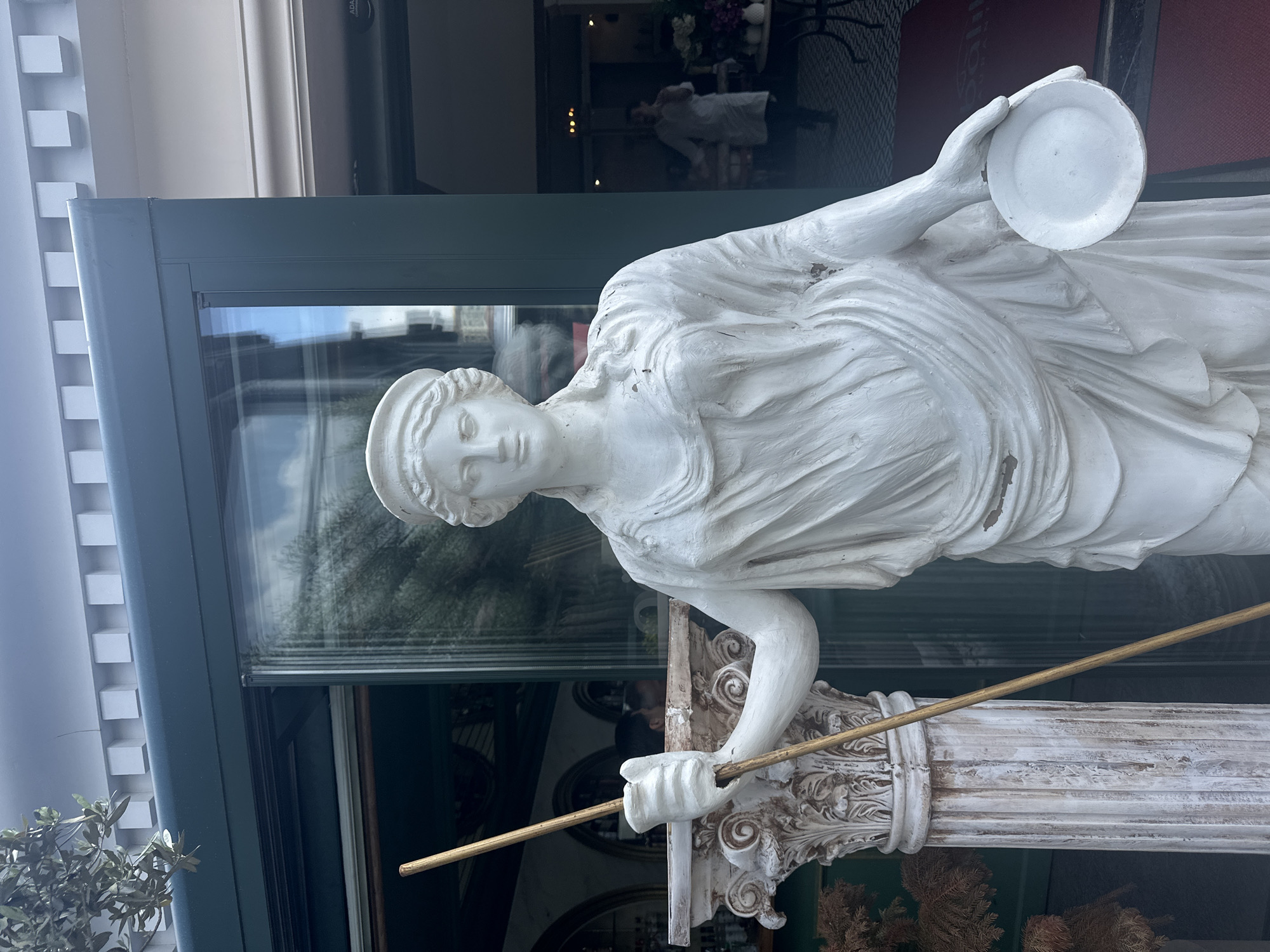 A vertical eye-level shot shows a white, stone or plaster sculpture of a Grecian woman holding a white plate aloft in her right hand and a golden-colored rod in her left hand. The statue stands in front of a large glass window of a building, with a grey or muted blue frame. The woman is wearing a draped garment. The statue appears to be in Başakşehir, Turkey. The interior of the building is visible through the window. Behind the statue, the pillar it is mounted on can be seen. The base of the pillar is visible with its decorative carvings.
