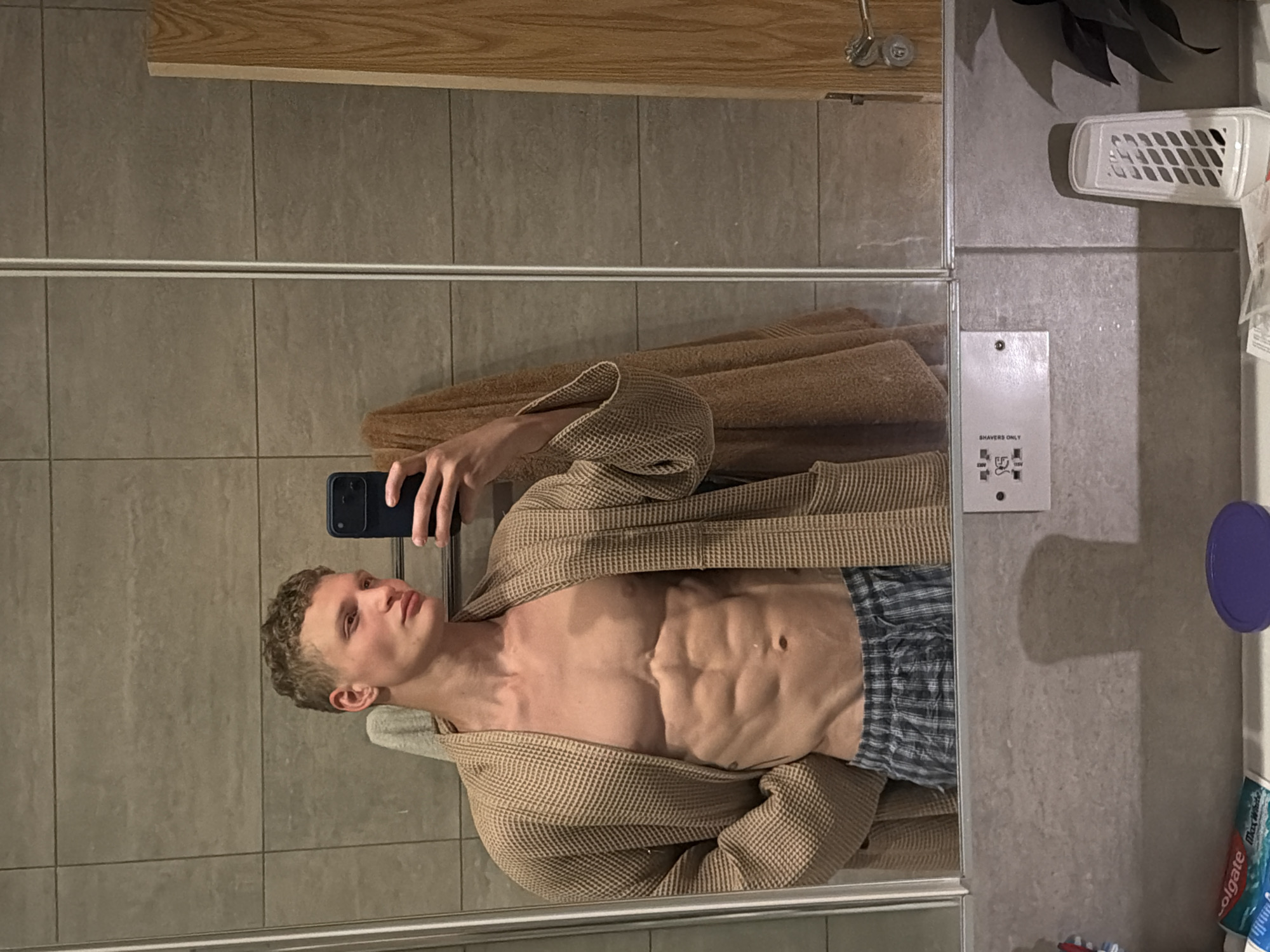 The image shows a reflection in a bathroom mirror. A light-skinned male with short, curly blond hair is holding a phone and taking a selfie. He is wearing a brown waffle-weave robe that is open in the front, revealing a muscular torso. He is also wearing checkered pajama pants. The bathroom has light gray tile walls and a light wood countertop. In the background, there is a white shaver socket, a white toiletries holder, a purple lid, and toothpaste on the sink. There is no information to suggest that the image was taken in London, United Kingdom.