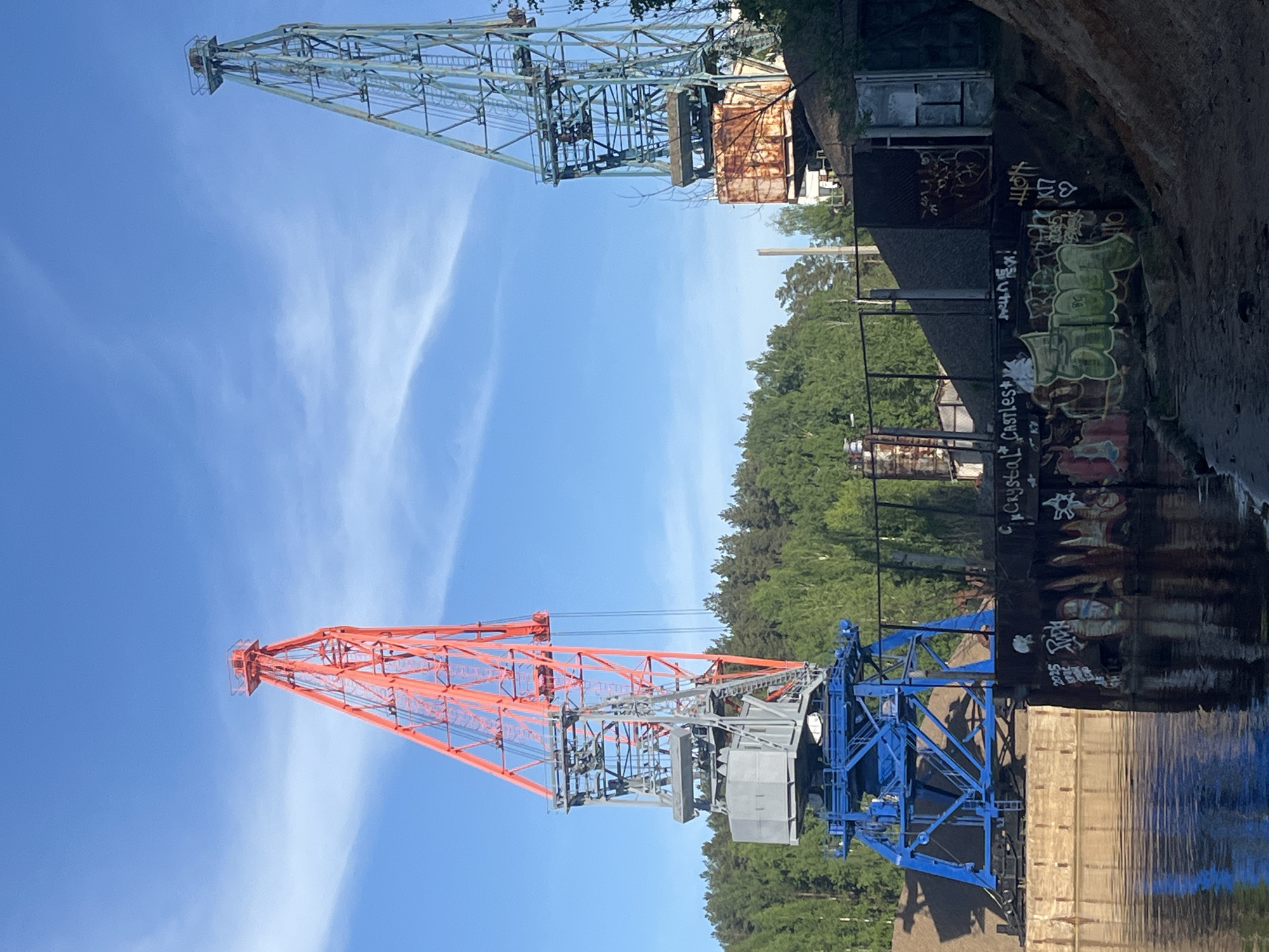 The image shows two cranes in the city of Chaikovsky, Russia. The crane on the right side of the image is painted in an orange hue while the one on the left is blue. In the background there is a body of water that seems like a river that reflects these cranes as well. The surrounding area has buildings with graffiti present, trees, and vegetation. The sky is mostly blue.