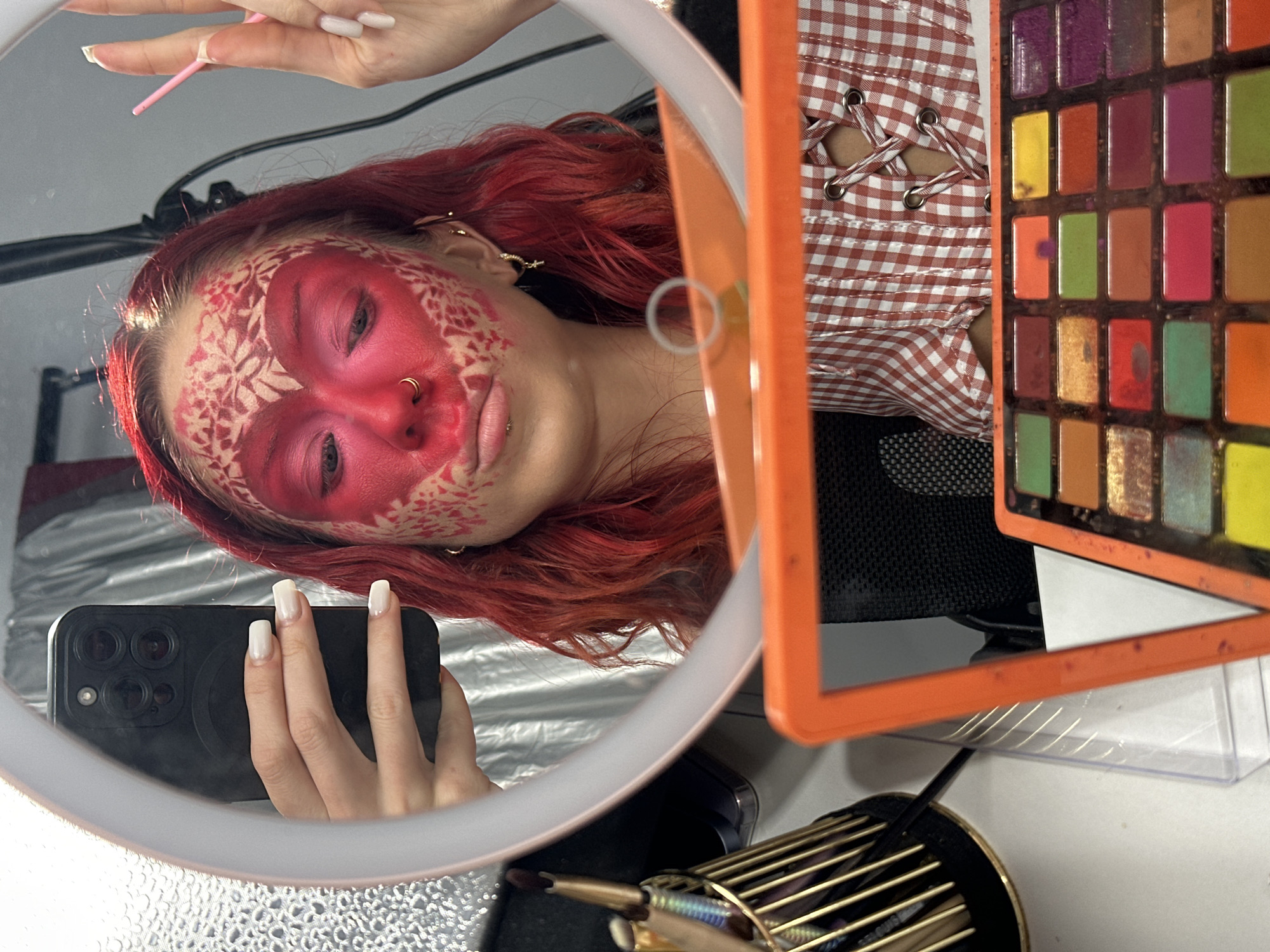 In a well-lit indoor setting, a young woman with vibrant red hair is applying makeup. She is looking into a ring light-equipped mirror, capturing a selfie with her phone. Her face is adorned with an intricate pattern of pink and white makeup, resembling a heart shape on her forehead and extending down her cheeks, with vine-like designs. She is wearing a red and white checkered top with lace-up detailing. In the foreground, a palette of colorful eyeshadows is visible. To the left, a collection of makeup brushes rests in a golden holder. The scene appears to be a personal vanity area, possibly in a home or a makeup studio. There is no indication of a specific location in North Kuta, Indonesia.