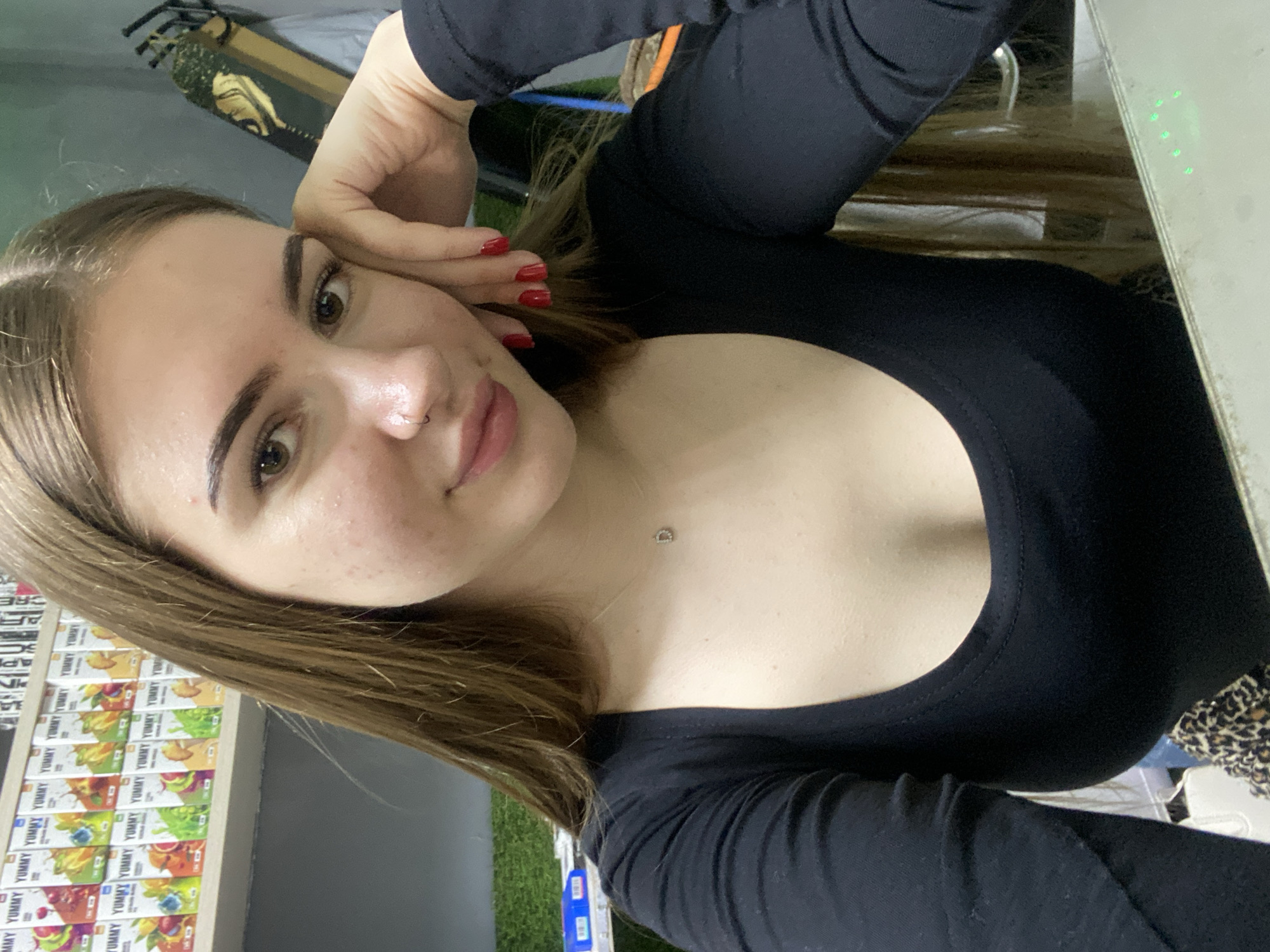 An angled, close-up selfie shows a young woman indoors. She has light brown hair, dark eyebrows, brown eyes, and a nose ring. Her lips are a soft pink, and she wears a black long-sleeved top with a wide, rounded neckline. A small silver necklace with a 'D' charm hangs on her chest. Her right hand, adorned with red nail polish, is gently resting near her face.
In the background to her left, a shelf displays multiple "YUMMY" branded juice boxes with various fruit illustrations, suggesting a retail or cafe setting. To her right, a surface featuring several small green lights is visible. Further back, what appears to be artificial grass or turf is present, along with a skateboard showing a design, possibly a Buddha head. In the upper left background, there is partially visible and indiscernible text on a wall or signage. The woman looks directly at the camera with a neutral to slightly content expression.