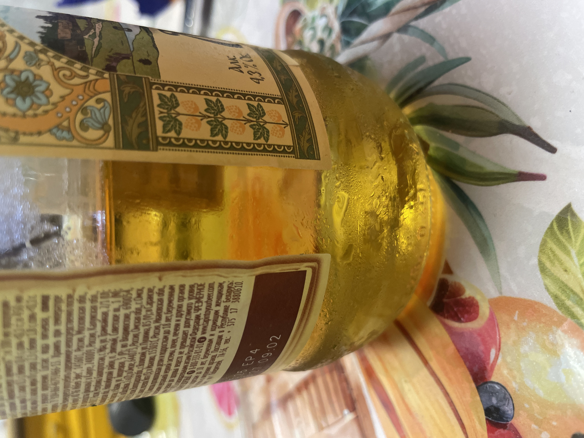 A close-up shot focuses on the lower portion of a glass bottle filled with a golden liquid. The bottle is placed on a surface adorned with a colorful pattern of fruits, possibly oranges and other citrus fruits, and green leaves. Condensation is visible on the exterior of the bottle, indicating it is chilled. A label on the bottle features text in Russian. One section displays "АЛК. 4,3% об." which translates to "ALCOHOL. 4.3% vol." Another part of the label shows the following text: "26 EP4 27 09:02." A larger portion of the label contains extensive text, which appears to be product information or warnings. A snippet from this text, visible in a cropped image, reads "Изготовитель и уполномоченная организация на принятие претензий от потребителей в Республике Беларусь: ОАО «Сталовары», ул. Пушкина, 85 (М-р Саранское, ул. Пушкина, 85) М-р Саранское, 690054, Россия, Смоленская обл., г. Орел, ул. Пушкина, 85 (М-р Саранское, 690054, Россия, Смоленская обл., г. Орел, ул. Пушкина, 85)." This text indicates manufacturers and authorized representatives for consumer claims in Belarus. It also mentions specific locations in Russia: Smolensk Oblast, Oryol, and a rural locality "Saranskoe". Additionally, the text mentions a phone number: "+375 17 3880610". The text further states: "Не превышает допустимого уровня. Пищевая ценность на 100г продукта: белки-0г, жиры-0г, углеводы-3,5г. Энергетическая ценность - 138 ккал (578 кДж). Состав: вода, солод ячменный, хмель. Не употреблять во время беременности, кормления грудью, управления транспортными средствами, а также лицам моложе 18 лет. Хранить в затемненном месте при температуре от 0 до 25°С. Срок годности 12 месяцев. Дата розлива указана на бутылке. Сделано в России." This translates to: "Does not exceed the permissible level. Nutritional value per 100g of product: proteins-0g, fats-0g, carbohydrates-3.5g. Energy value - 138 kcal (578 kJ). Ingredients: water, barley malt, hops. Do not consume during pregnancy, breastfeeding, while operating vehicles, or by persons under 18 years of age. Store in a shaded place at a temperature from 0 to 25°C. Shelf life 12 months. Bottling date is indicated on the bottle. Made in Russia." While the image itself does not provide specific details about the city of Laishevo, Russia, the Russian text on the label suggests that the product is manufactured or distributed within Russia and potentially exported to Belarus. The mention of "Смоленская обл., г. Орел" indicates a manufacturing location within Russia. Without additional context or geographical markers within the image, the precise location of Laishevo cannot be