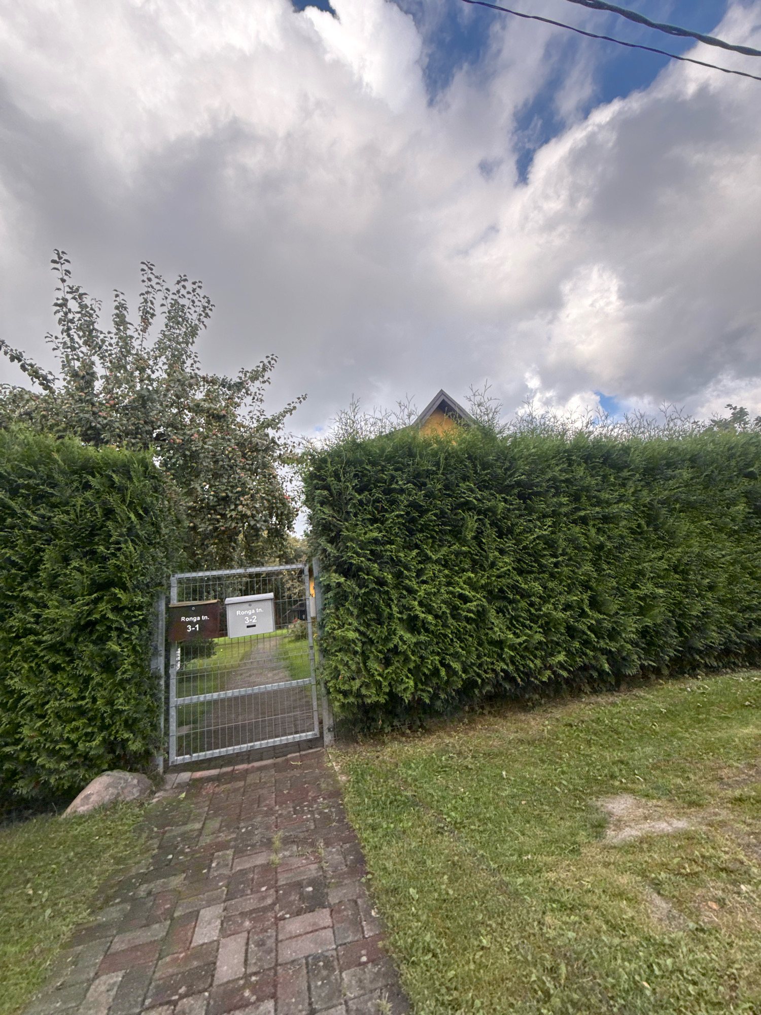 The image captures an outdoor scene featuring a fenced entrance to a property. The fence is made of metal and has two mailboxes attached. One mailbox is labeled "Ronga tn. 3-1" and the other is labeled "Ronga tn. 3-2". Beyond the fence, a gravel path leads to a house partially visible with a yellow roof. The property is bordered by neatly trimmed green hedges and a large tree with fruit. The ground is covered with green grass and a brick pathway. The sky is cloudy with patches of blue. The location is Ronga Street in Tallinn, Estonia.