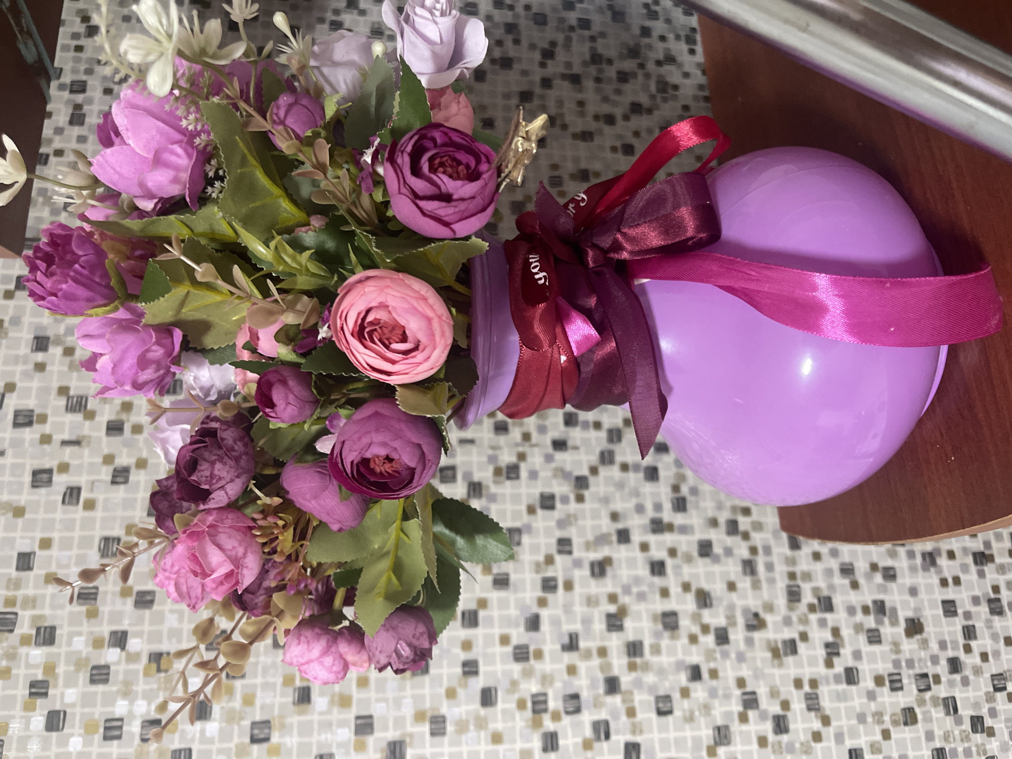 A bouquet of artificial flowers, predominantly in shades of pink and purple, is displayed in a light purple, round vase. The vase is adorned with dark red and pink ribbons tied in a bow, with one ribbon featuring white text that appears to say "you". The background is a mosaic tile wall with a pattern of small, square tiles in varying shades of grey, beige, and gold. The scene is set on a dark wooden surface. The location is specified as Марковский, Russia. There are no people or animals in the image.