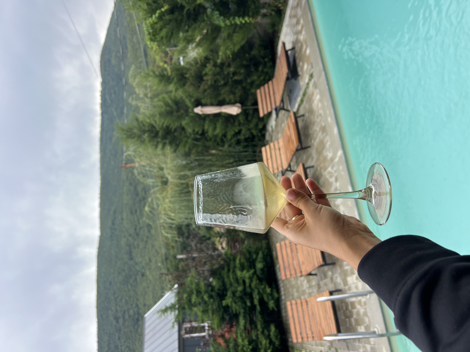 A person's hand, adorned with a pink-nailed finger and a ring, holds a clear wine glass filled with a pale yellow liquid, positioned in the foreground against a backdrop of a swimming pool and a lush, green hillside. The swimming pool, filled with turquoise water, is visible in the lower right quadrant of the image, with its edge and pool ladder in the lower left. Several wooden lounge chairs are arranged on a stone patio behind the pool. In the middle ground, tall, dark green coniferous trees and a small building with a gray, sloped roof are visible. A light-colored umbrella stands behind the lounge chairs. The background features a densely forested, sloping hill under an overcast sky. The scene appears to be outdoors, likely at a resort or a private residence with recreational facilities, in the vicinity of Tsodoreti, Georgia, given the presence of a hillside and greenery. The overall atmosphere suggests a relaxed, possibly cool or rainy, day.