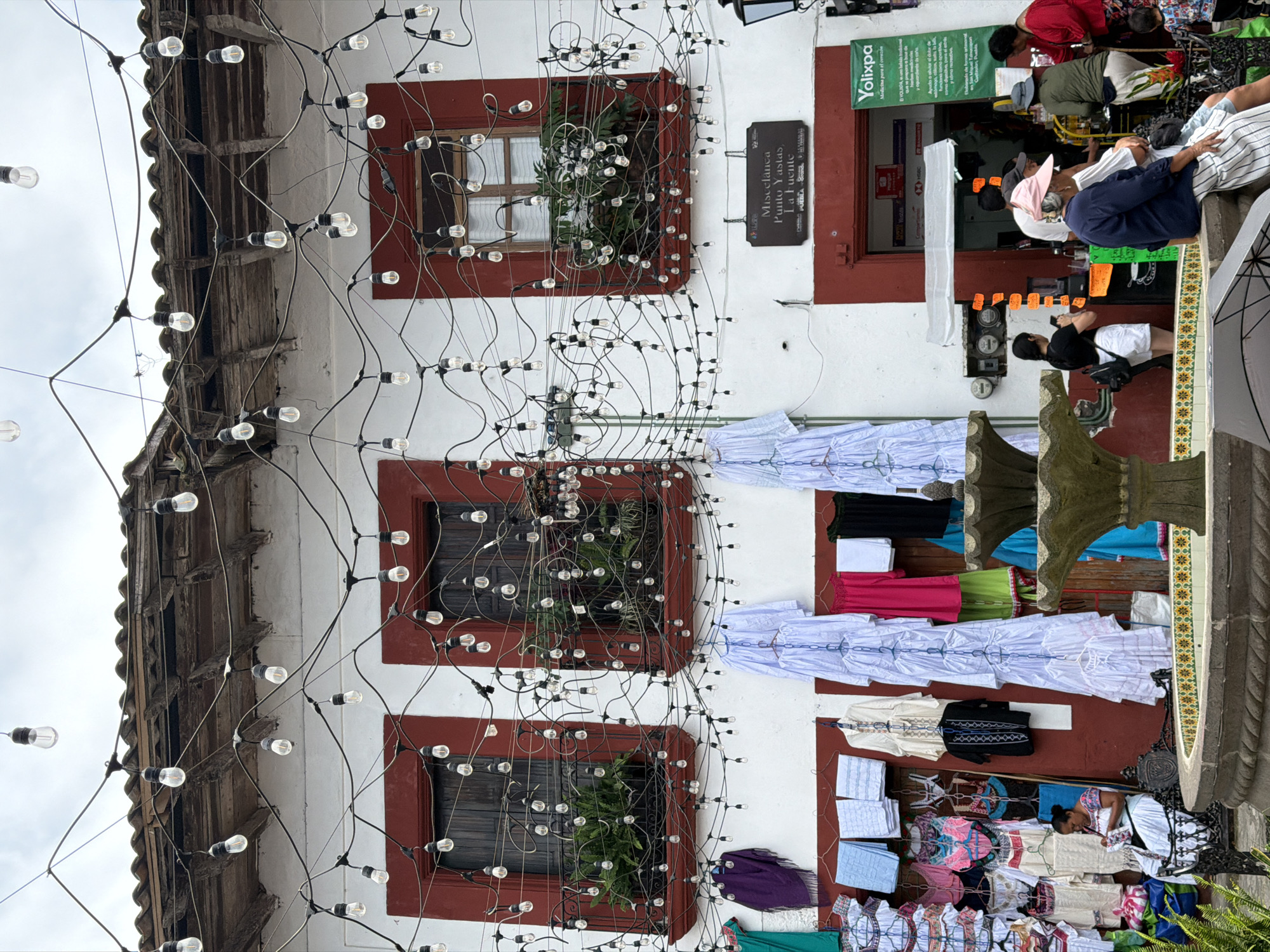 Here is a detailed description of the image:
The image captures a vertical view of a building facade adorned with a complex arrangement of string lights, which create a crisscross pattern across the building's exterior. The building has a white-painted exterior with dark brown window frames, and potted plants are visible inside some of the windows. Clothes are hanging from the building's exterior, possibly items for sale.
On the right side of the image, there are people seated and standing near a store named "Yolixpa" with an address and a logo that states, "Medicina para el corazón." There is a sign next to one of the windows, identifying the location as "Miscelanea Punto Yastas, La Fuente PUEBLA."
The image appears to be taken in Cuetzalan, Mexico, a location well-known for its historic architecture, indigenous communities, and traditional markets.