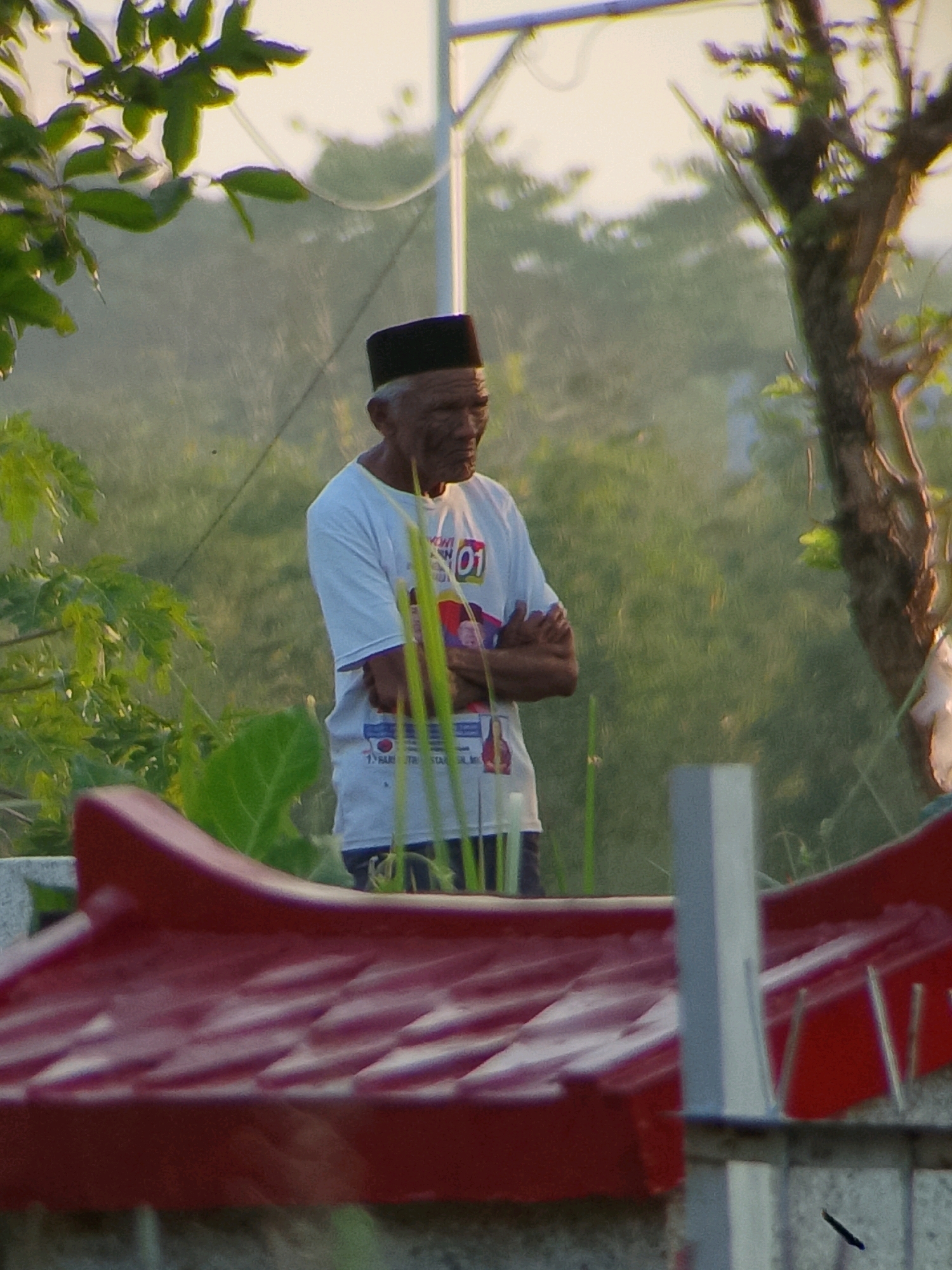 In Jember, Indonesia, an elderly man wearing a black cap and a white t-shirt stands with his arms crossed. The t-shirt has a graphic print on it. He is positioned outdoors, with lush green foliage and trees visible in the background, suggesting a natural or park-like setting. The lighting indicates it may be daytime, possibly late afternoon given the warm tones. In the foreground, a portion of a red structure, possibly a roof or a decorative element, is visible. The overall scene appears calm and quiet.