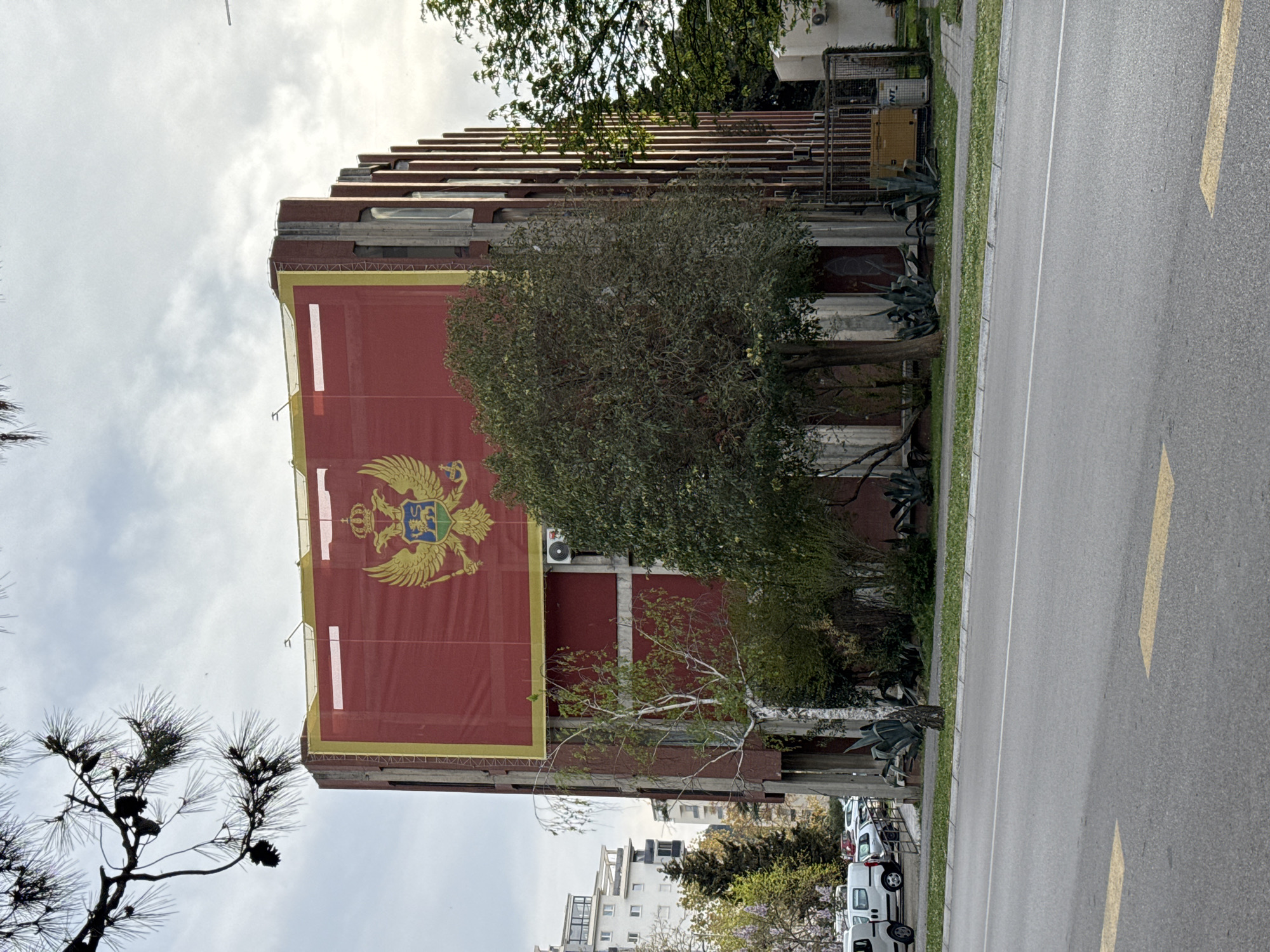 The image shows a large building in Bar, Montenegro, adorned with a massive banner displaying the national flag. The building's architecture is tiered, and it is set back from the street behind some trees and greenery. The street is paved and features yellow lane markings. Cars are parked along the street. The sky is overcast with diffuse clouds.