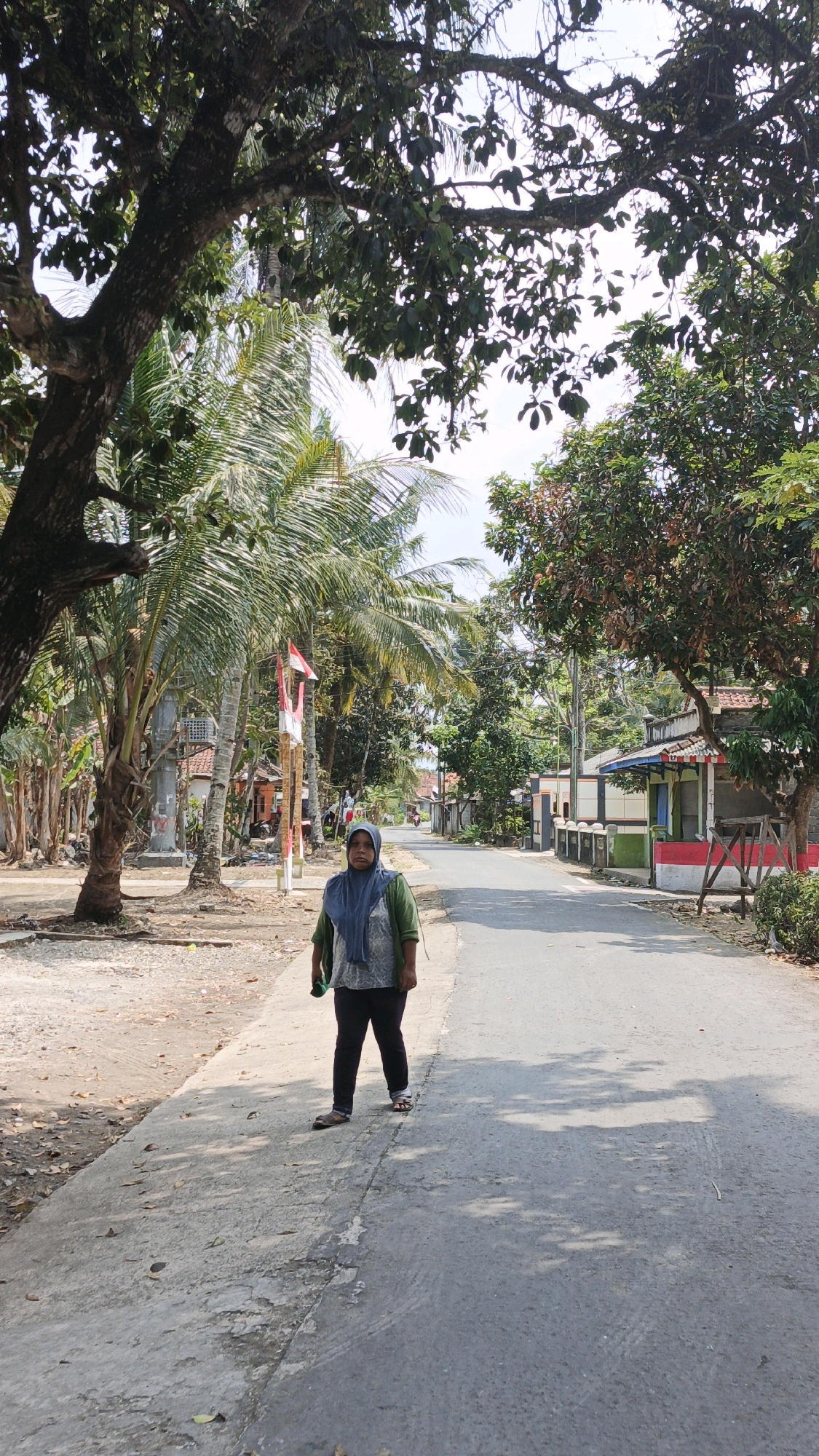In Cibenda, Indonesia, a woman stands on a paved sidewalk next to a road. She is wearing a green hijab and a long-sleeved green shirt over dark pants and sandals. She is facing the camera, holding a dark object in her right hand. The road is lined with palm trees and some buildings, one of which has a red and white facade. A Indonesian flag is visible on a pole near the woman. The scene is under a canopy of trees with dappled sunlight on the ground.