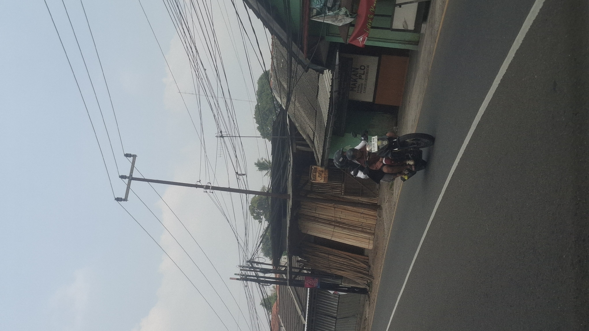 The image shows a street scene in Cilaku, Indonesia, on a cloudy day. A motorcycle carrying two people, a driver and a passenger, is traveling on the right side of the asphalt road. The motorcycle has a license plate with the inscription "B-3685 YNZ". To the right of the road, there is a building with a green facade and a sign that reads "MAKAN PLO". In front of the building, stacks of lumber are visible, leaning against a structure with a corrugated metal roof. Another building with a similar roof and a metal fence is located further to the left. Overhead, numerous power lines crisscross the sky, supported by utility poles. The environment appears to be a semi-rural or commercial area with a mix of residential and business structures.