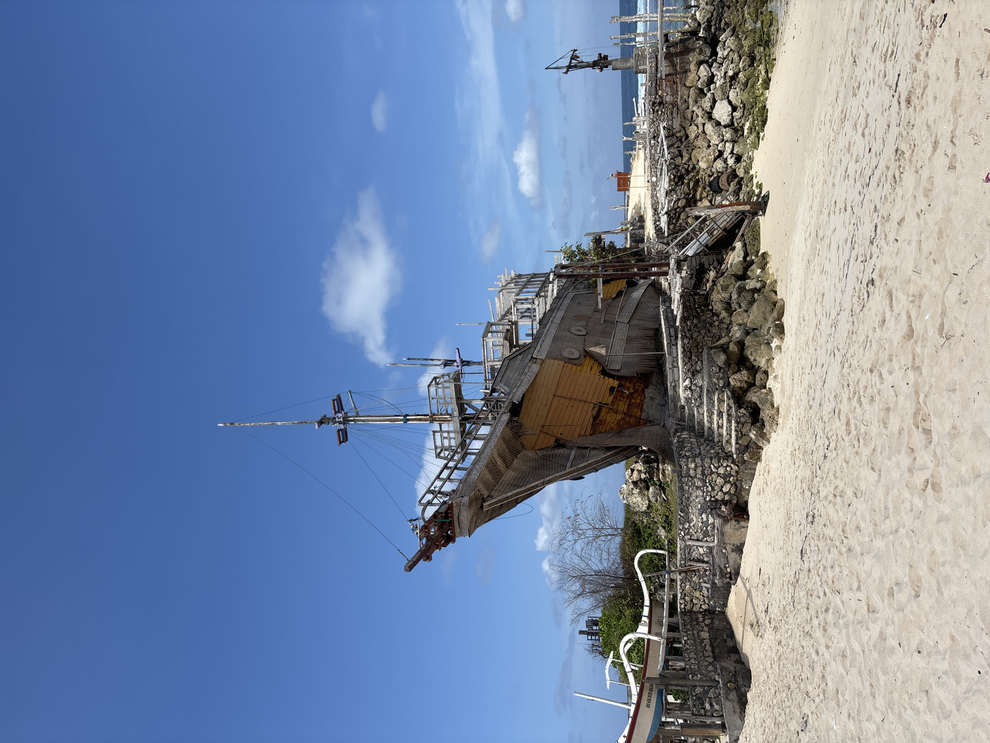 A wooden, unfinished replica of a pirate ship is positioned on a sandy beach in Kuta Selatan, Indonesia. The ship is beached and appears to be a static structure rather than a functional vessel. It has a tall mast, wooden decks, and a decorative bowsprit resembling a dragon's head. The ship is partially supported by a stone and wooden retaining wall, with stairs leading up to its structure. The beach is composed of light-colored sand. In the background, the ocean is visible under a bright blue sky with scattered white clouds. There are several other smaller, traditional Indonesian boats, known as jukung, pulled up onto the beach near the main structure. The surrounding area includes some rocky outcrops and sparse vegetation. No people are visible in the frame.