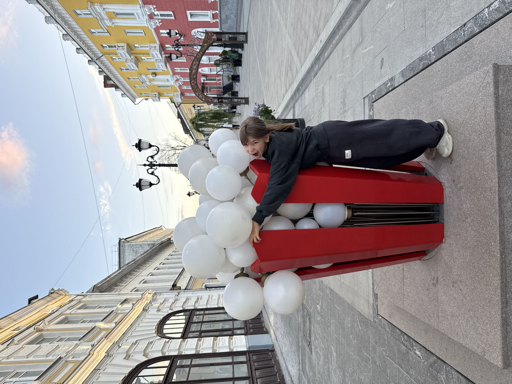 A young girl with brown hair is embracing a large, red, popcorn-shaped sculpture filled with white balloons. She is wearing a dark gray sweatshirt and dark gray sweatpants, and white sneakers. The sculpture is located on a paved street in what appears to be a European city. In the background, there are buildings with colorful facades, streetlights, and trees. The sky is a pale blue with some wispy clouds. The scene suggests a playful and festive atmosphere.