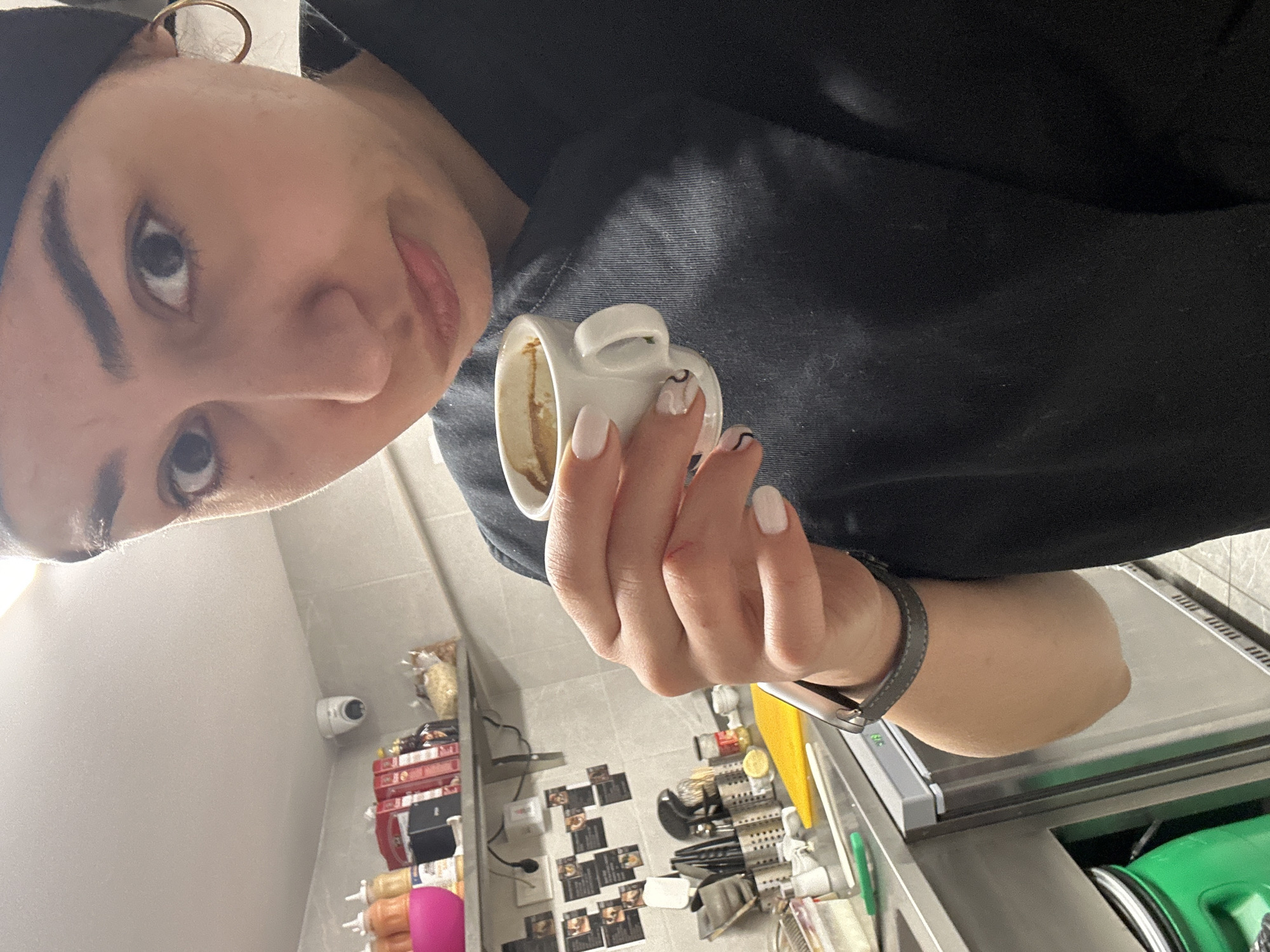 A woman is holding a small, white coffee cup with dregs visible inside. She is wearing a black shirt and a gold hoop earring. Her fingernails are painted white with black swirls. She has a smartwatch on her left wrist. The background shows a commercial kitchen environment with stainless steel counters, shelves stocked with food items, a vacuum sealing machine, and a CCTV camera mounted on the wall. Framed black and white photos are mounted on the wall below the shelves. I cannot determine if this photo was taken in Burgas, Bulgaria.