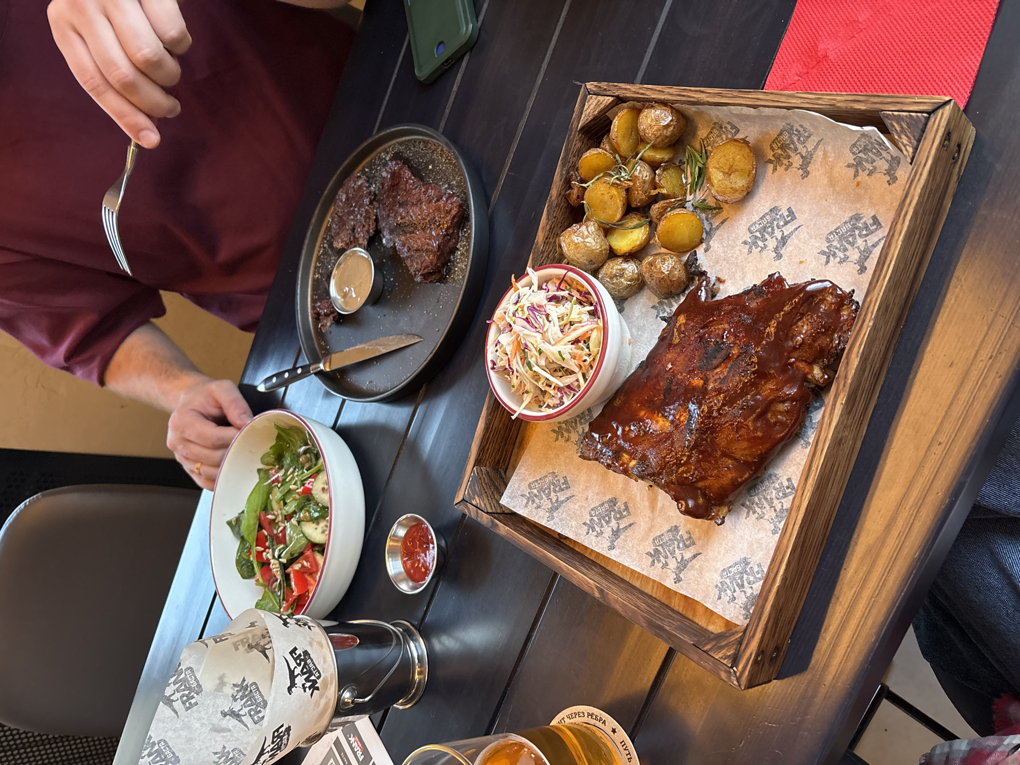 The image shows a table in a restaurant in Krasnoyarsk, Russia, laden with food. A person with a fork is about to eat a steak from a plate that also contains a small bowl of sauce and a knife. A wooden tray holds ribs, roasted potatoes with rosemary, and coleslaw. A bowl of salad, a small dish of red sauce, and a beer glass in an ice bucket are also on the table. The table is dark and possibly made of wood. The overall setting suggests a casual dining experience.