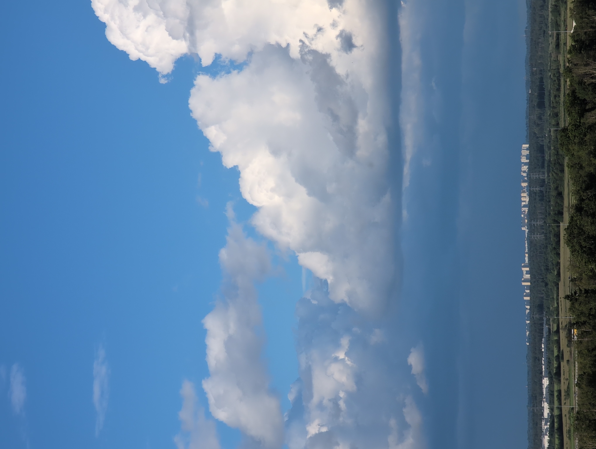 The image shows a bright blue sky with scattered white and gray clouds. The clouds are dense and puffy, with some areas appearing darker, suggesting possible rain or storm clouds. A thin, elongated cloud stretches across the upper right portion of the frame, obscuring the sky behind it.
In the distance, a treeline forms a dark green band along the horizon. Beyond the trees, a row of white, multi-story buildings is visible, suggesting an urban or suburban environment. The buildings are lined up in a straight formation, with some electrical poles or towers interspersed among them. The overall scene depicts a transition from a cloudy sky to a developed landscape.
The lighting suggests it is daytime, likely midday or afternoon, given the brightness of the sky. The weather appears to be mixed, with sunshine and clouds. The presence of buildings and infrastructure implies a populated area, but no people are visible in the image. The composition is dominated by the sky, with the landscape occupying a smaller portion of the frame at the bottom. No text is visible in the image.