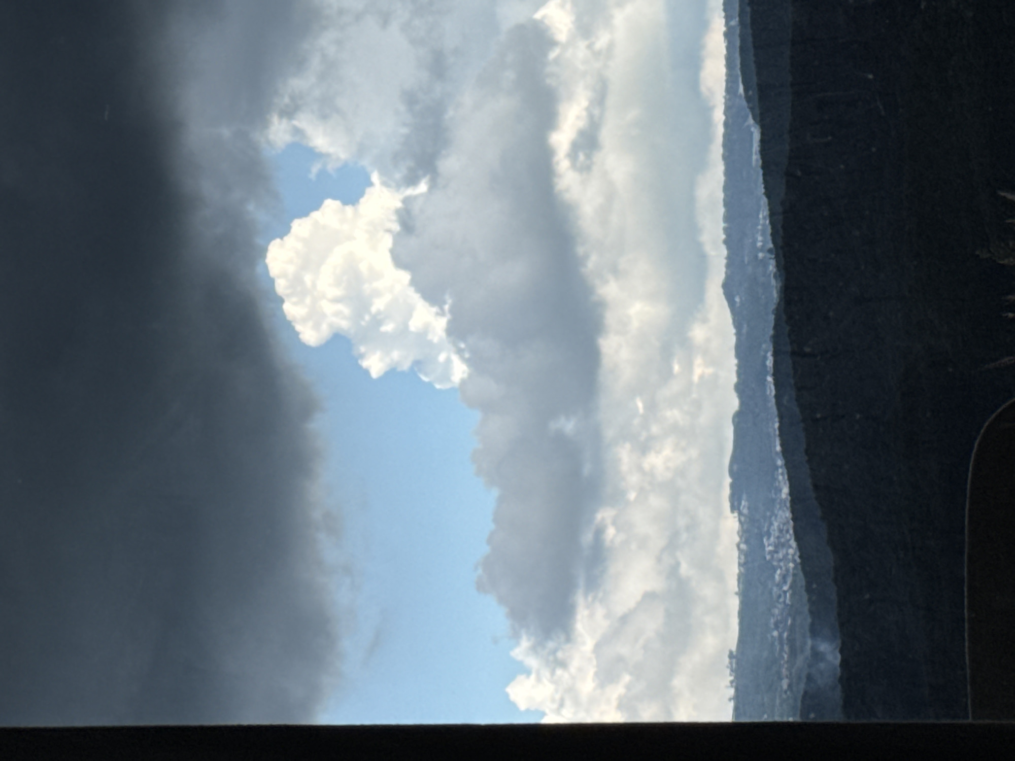 A photograph captures a panoramic view of the city of Đa Lộc, Vietnam, dominated by dramatic cloud formations and mountainous terrain. The foreground is defined by the dark, silhouetted forms of rolling hills, their dense foliage obscuring any ground-level details. These hills create a stark contrast against the sky, which is filled with a dynamic interplay of light and shadow.
The sky is a prominent feature, with vast expanses of both dark, ominous storm clouds and bright, fluffy cumulus clouds. The storm clouds, particularly in the upper left quadrant, appear heavy and laden, suggesting an approaching or receding weather system. In stark contrast, a large, cauliflower-like white cloud formation stands out against a patch of bright blue sky in the upper center of the image. This bright cloud is illuminated from above, indicating sunlight breaking through or a gap in the darker cloud cover. Below these, a layer of lighter, scattered clouds stretches across the horizon.
In the mid-ground, a town or city is visible nestled within the undulating landscape. Numerous buildings, appearing as a cluster of small structures, are scattered across a valley or plateau. The density of these buildings suggests a populated area. A faint wisp of smoke or steam rises from one of the darker hills in the mid-ground, possibly indicating localized activity or atmospheric conditions.
The overall impression is one of natural grandeur, with the weather and landscape taking center stage. The absence of any visible people, vehicles, or specific man-made structures in the immediate foreground or mid-ground, beyond the distant settlement, keeps the focus firmly on the environment. The image is shot from an elevated perspective, looking out over the landscape and towards the horizon, with a dark, vertical element on the left edge of the frame suggesting it was taken from within a structure or through a narrow opening.