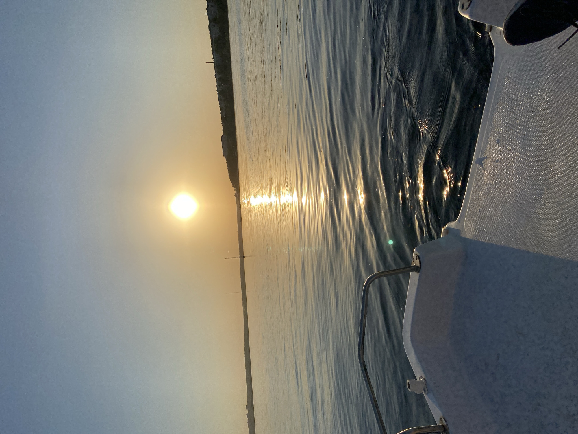 The image shows a sunrise over water, likely a lake or river. The sun is low on the horizon, casting a golden light across the water. Part of a boat is visible in the foreground, with a metal railing and a white, textured surface. On the opposite shore, a building or city skyline is visible in the distance. The image does not appear to be in or near Izhevsk, Russia.