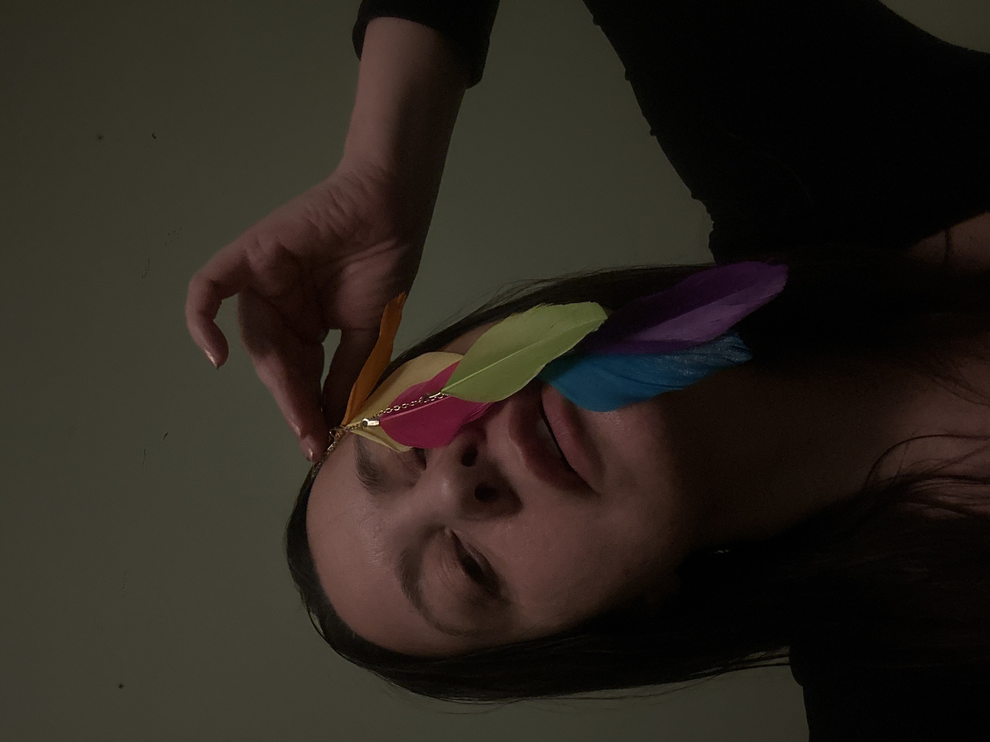 A close-up, slightly rotated studio shot depicts a woman with closed eyes, adorned with a colorful feathered headpiece. The feathers are arranged in a spectrum of colors, including orange, pink, yellow, blue, and purple. A golden chain is attached to the base of the feathers. A hand is poised just above the woman's forehead, near the feathers. The woman has dark hair and is wearing a black garment. The background is a muted, neutral tone. No information indicates the location in Riga, Latvia.