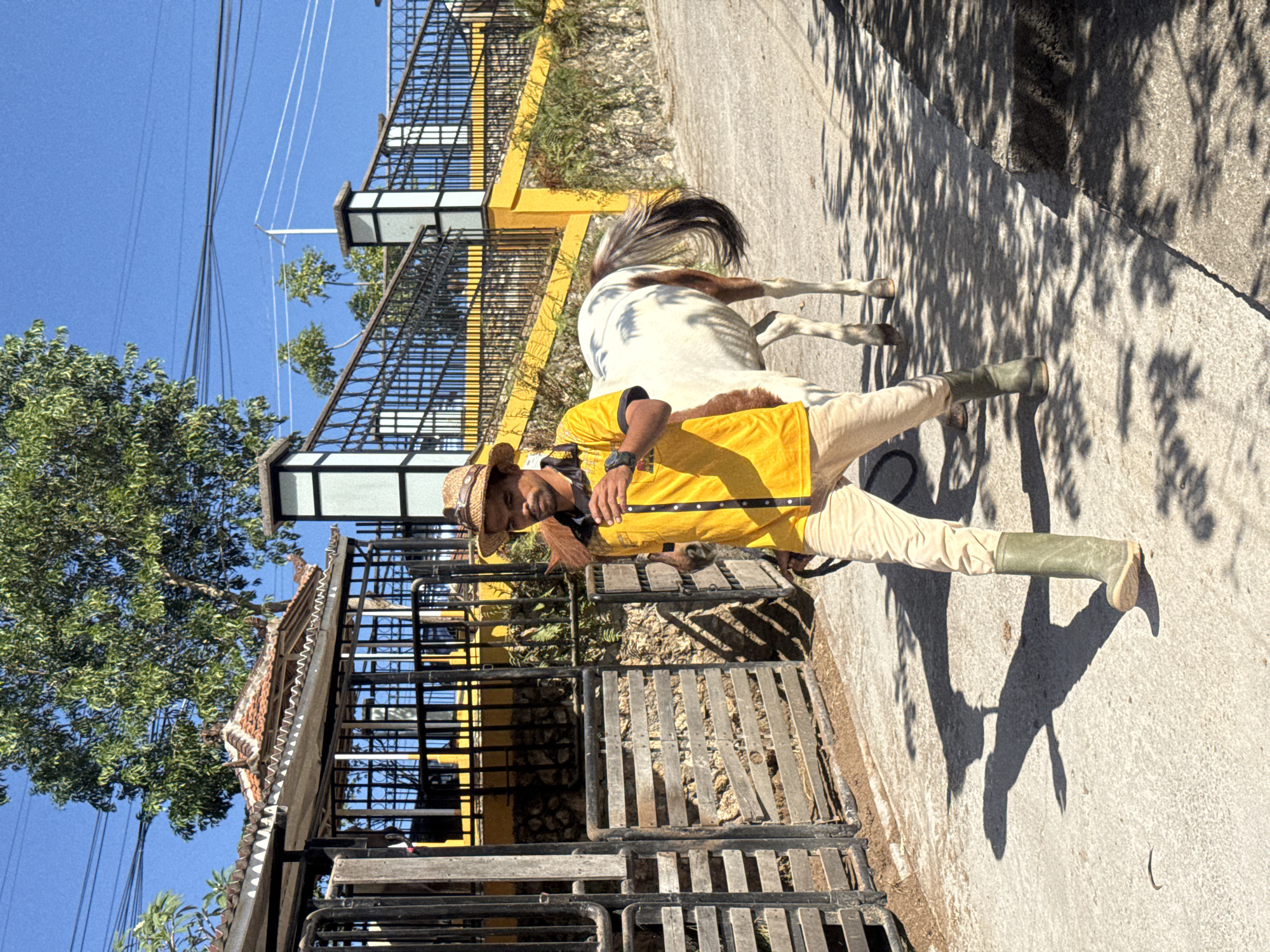 A person wearing a straw hat, a yellow shirt with black trim, beige pants, and green boots walks along a paved path next to a white and brown horse. The person is holding a lead rope attached to the horse. The path is on a slight incline, and there are shadows of trees on the ground. In the background, there is a black metal fence with yellow accents, and beyond that, a building with a tiled roof and greenery. The scene appears to be outdoors in a sunny environment. The style of the building and the surrounding vegetation suggest a location in Bali, potentially Kuta Selatan, Indonesia, given the provided context.
