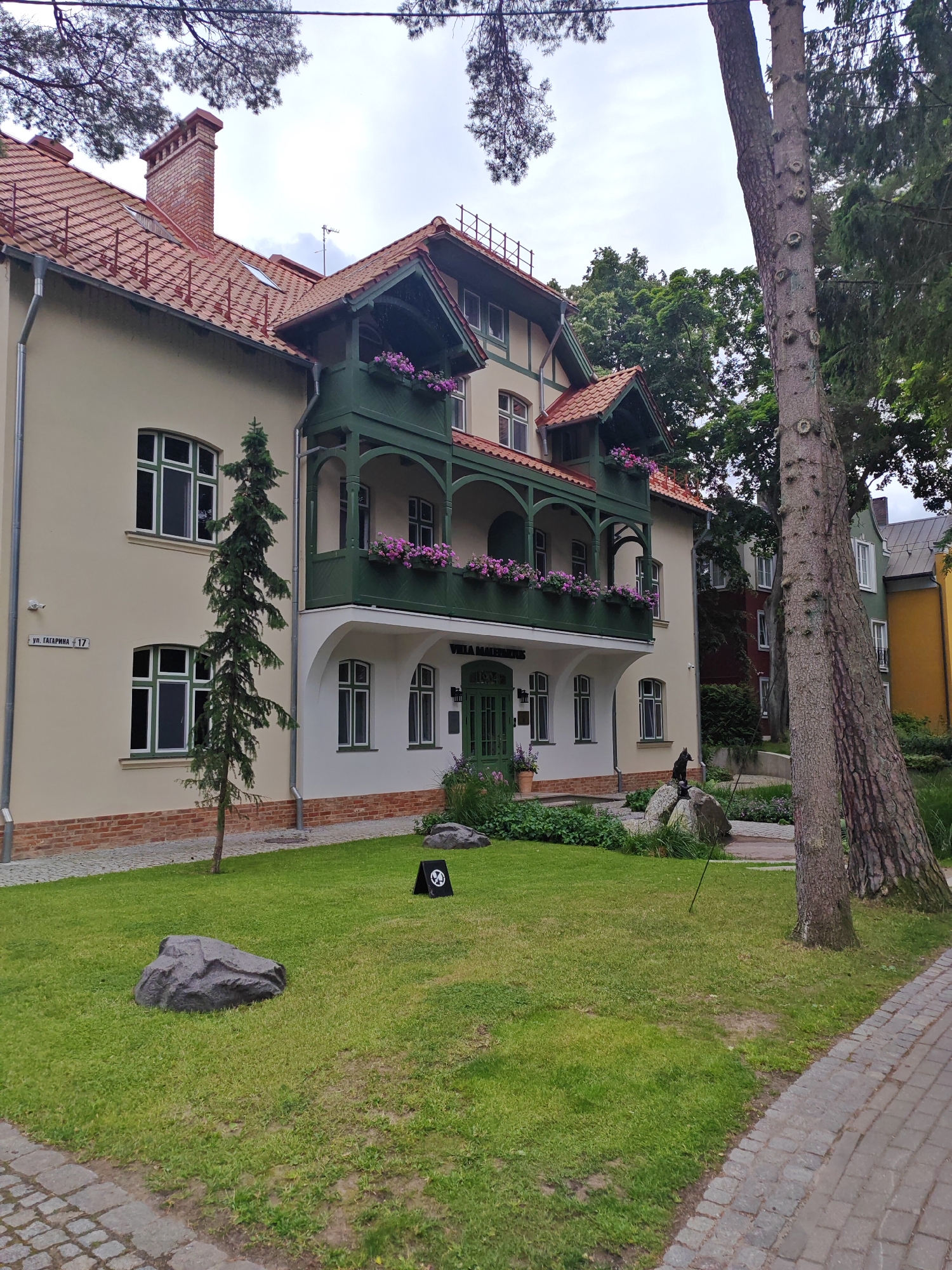 A multi-story building with a red tile roof is situated in a grassy area with trees. The building is light yellow with green trim around the windows and a green balcony adorned with pink flowers. A sign on the building reads "VILLA MARINAIS" and another sign on the side of the building indicates "ул. Гагарина 17." In front of the building, there are several large rocks and a small evergreen tree. A paved walkway runs along the right side of the scene, bordered by grass and trees. A black dog sculpture is perched on a rock in front of the building. In the background, other buildings with different colored facades are visible. The sky is overcast. The overall scene depicts a residential or hotel property in Svetlogorsk, Russia.