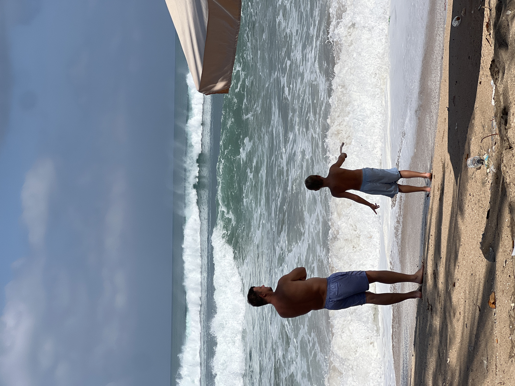Two individuals, an adult male and a child, stand on the sandy shore of Kuta Beach, Bali, Indonesia, facing a turbulent ocean with large waves. The man, with his back to the camera, wears blue striped shorts and has his arms crossed. The child, also facing the ocean, is dressed in light blue shorts and holds a stick. The water is a deep turquoise, with white foam from the crashing waves. In the background, multiple lines of waves are visible, and the sky is overcast with scattered clouds. A large, tan umbrella is partially visible in the upper right corner of the frame. The sand in the foreground is littered with debris, including plastic bottles and leaves, indicating potential environmental concerns on the beach. The scene captures a moment of observation of the powerful ocean on a cloudy day in Kuta.